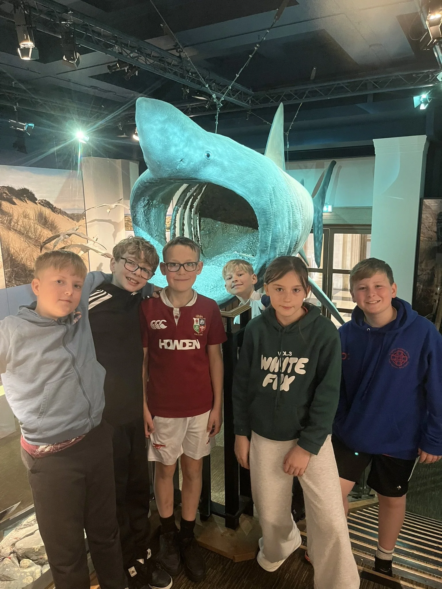 A group of six children standing in front of a large model of a prehistoric whale inside a museum. The model is blue-gray with an open mouth, displaying teeth, and a dorsal fin. The children are smiling and posing for the photo.