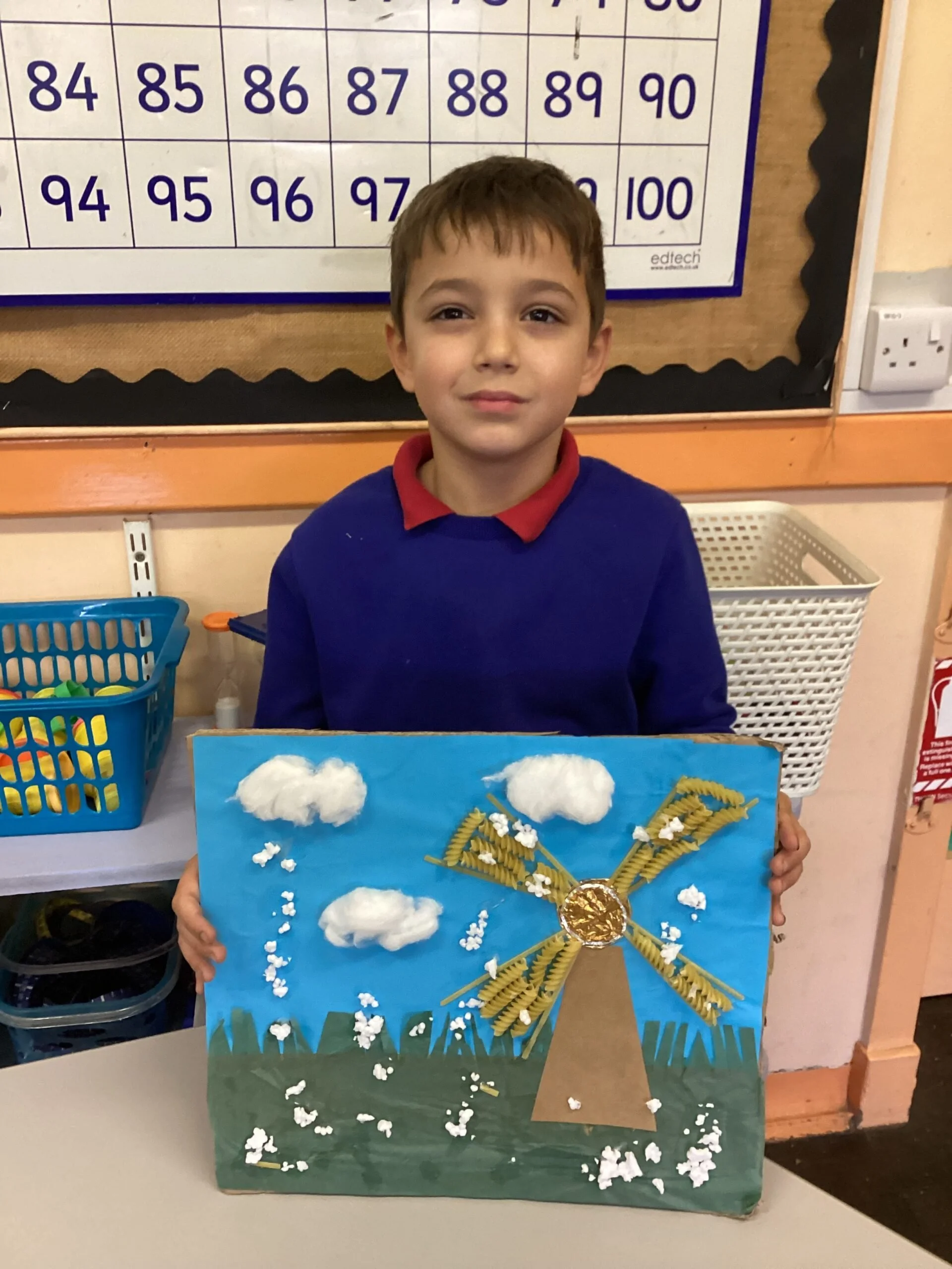 A young boy in school uniform holding a colorful craft project featuring a blue background, white cotton clouds, and golden pasta arranged as a sun with radiating rays. He is standing in classroom.