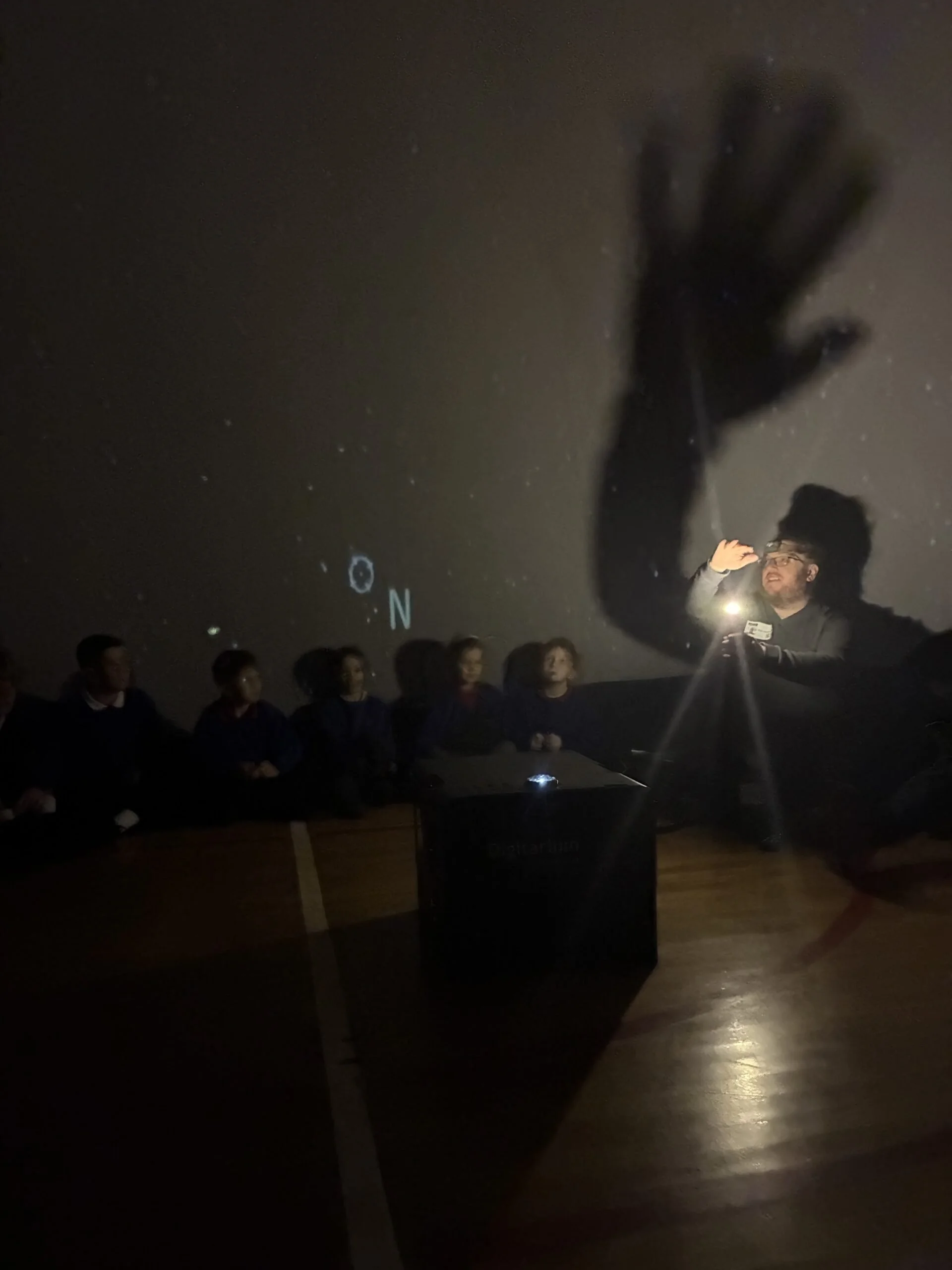 A person holding a flashlight in front of a dark wall, with a shadow of a hand projected onto the wall and a group of people watching.