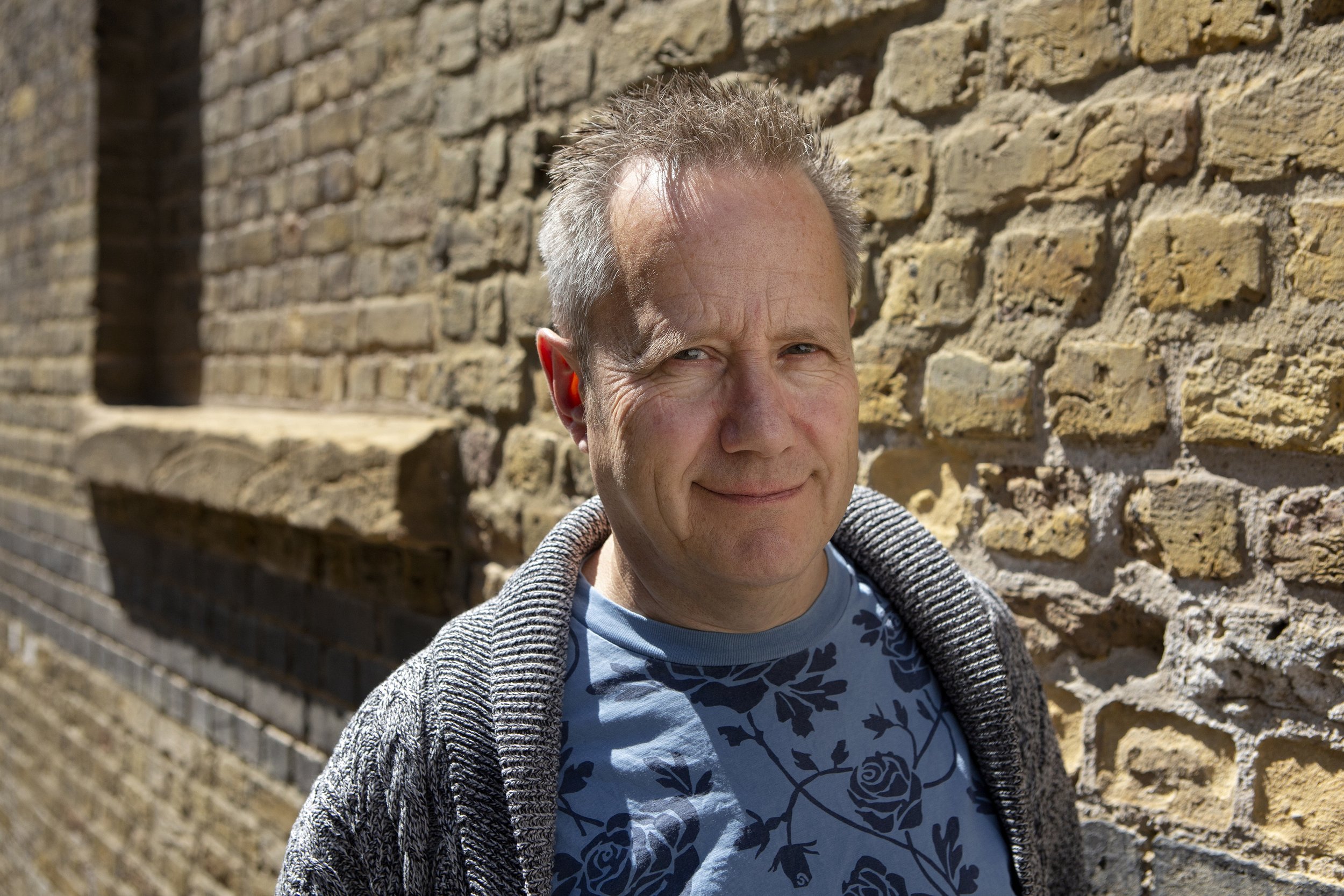 A middle-aged man with blond-grey hair smiling outdoors next to a brick wall.