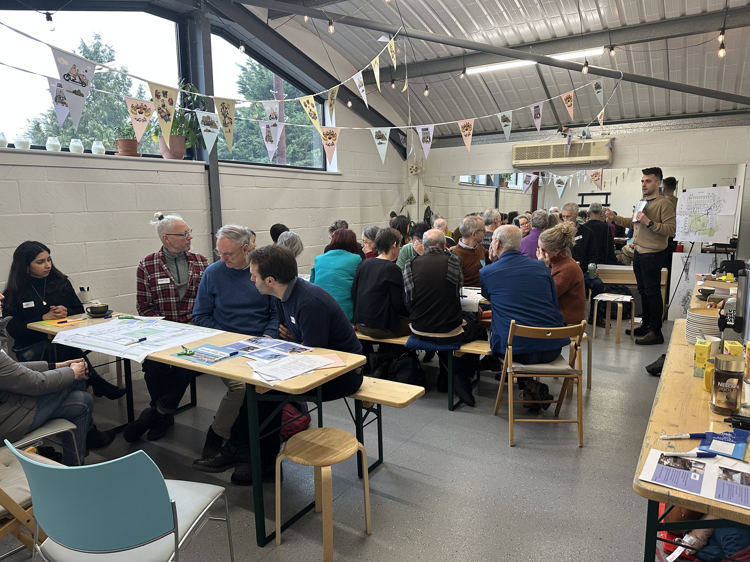A large group of people gathered indoors at tables, engaged in discussions, with some taking notes. Decorations include hanging bunting with illustrations. A person stands at the front holding a chart and addressing the crowd. The room has large windows, plant pots on the windowsill, with a casual and collaborative atmosphere.