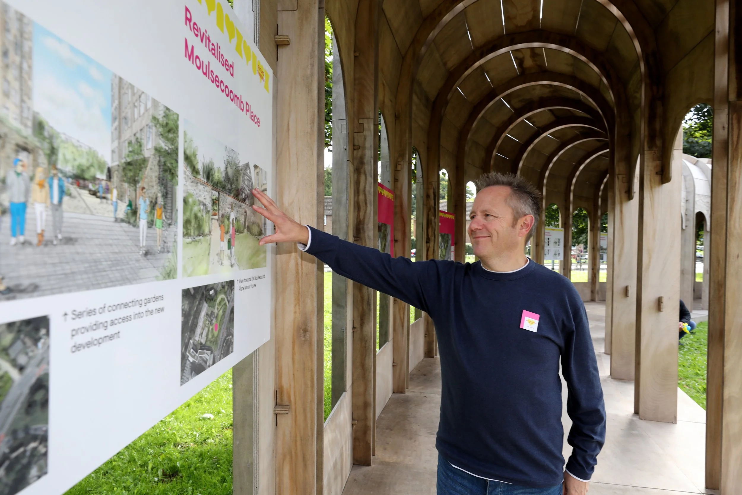 A man examining an information board at an outdoor exhibition under a wooden pergola. The board displays an illustration of a street scene with people and buildings, and contains text about revitalised Moulsecoomb Place.