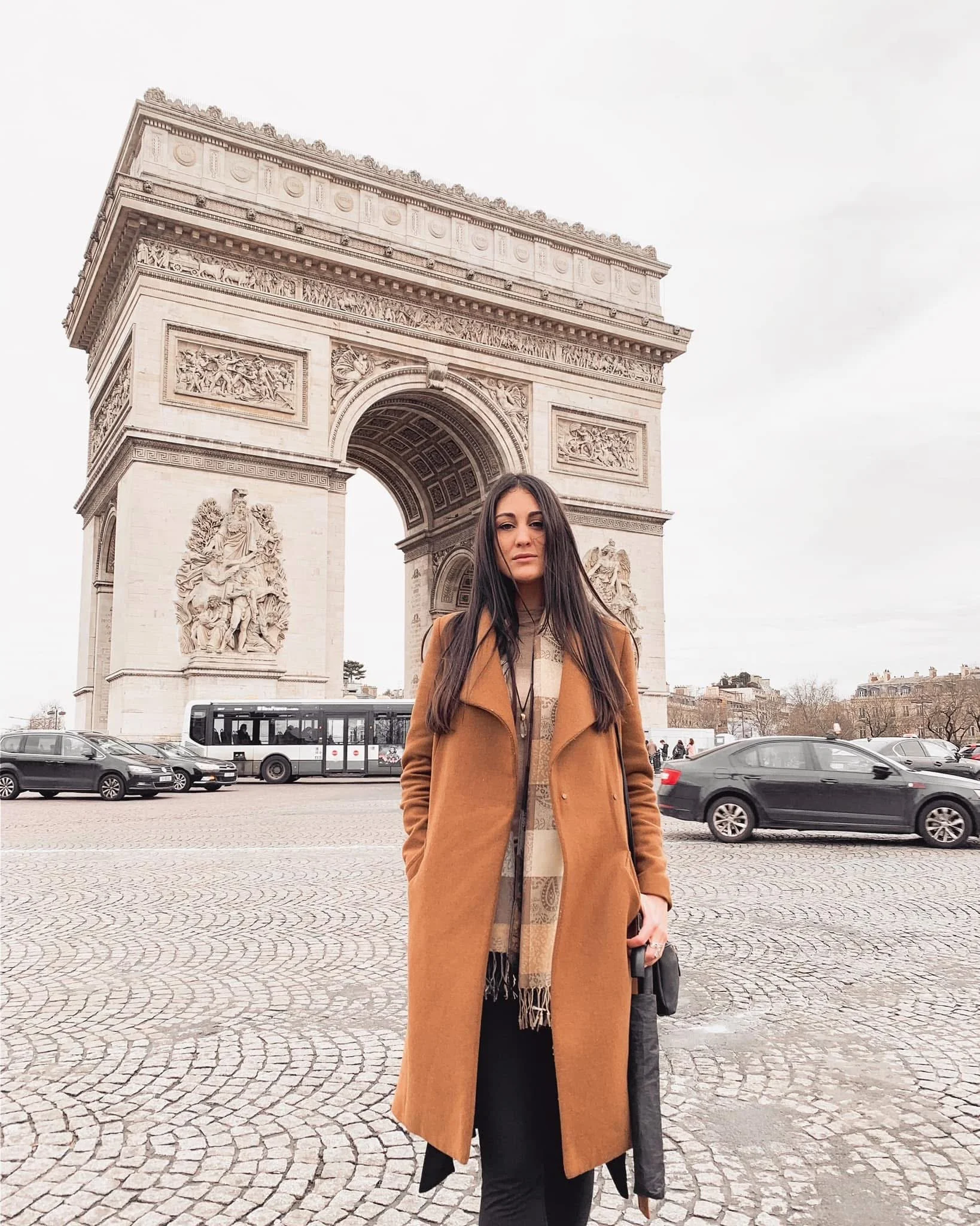 A woman with long dark hair wearing a camel-colored coat and patterned scarf standing on cobblestone ground in front of Arc de Triomphe in Paris, France, with cars and people in the background under an overcast sky.