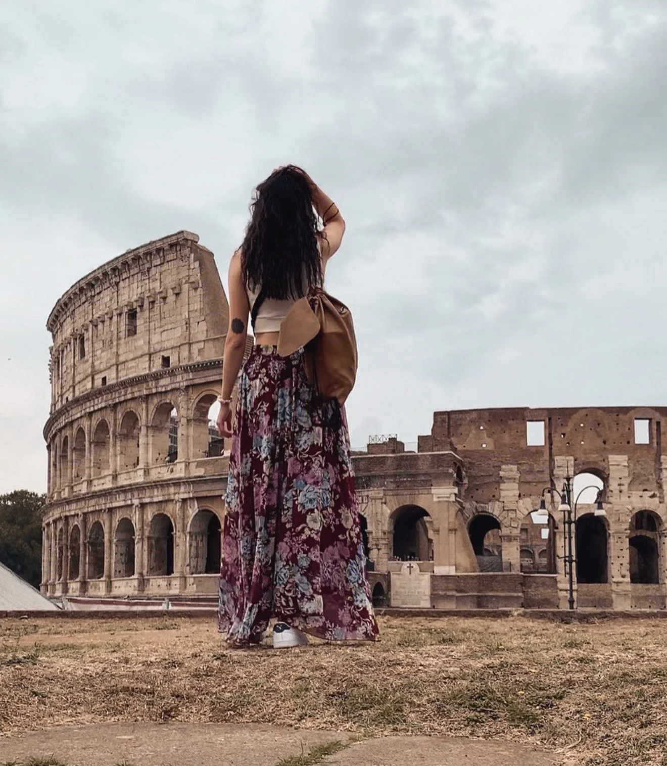 A woman with dark hair, wearing a floral skirt, a beige crop top, and white sneakers, standing in front of the Colosseum in Rome, Italy.