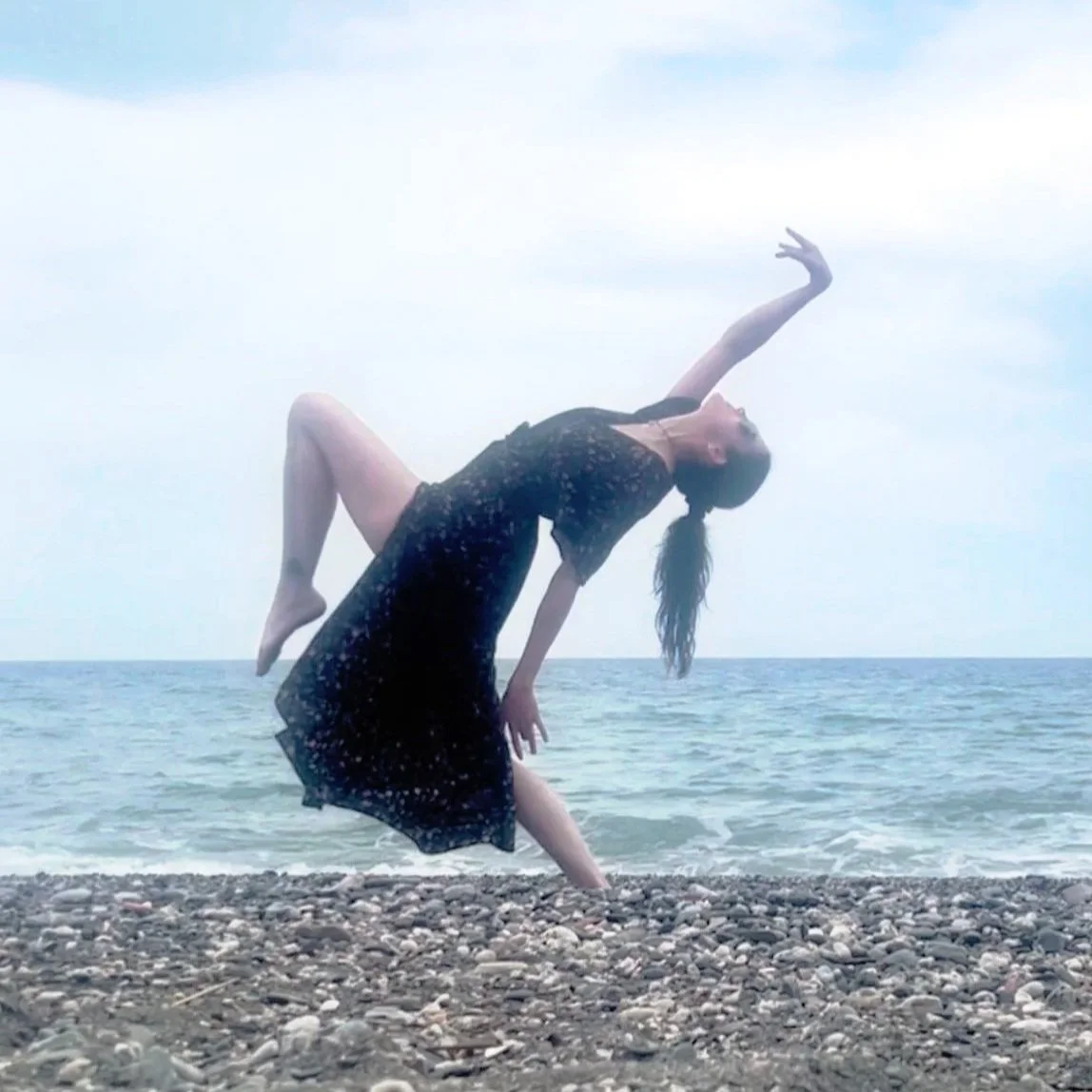 A woman wearing a black dress props her leg and arm while standing on a pebble beach near the ocean, with a cloudy sky above.