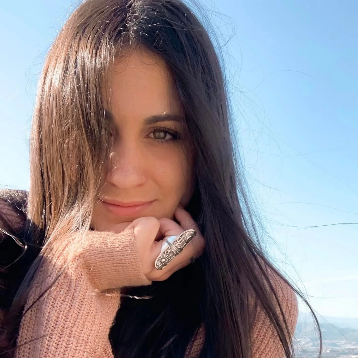A young woman with long dark hair and light colored eyes is smiling softly at the camera. She is wearing a ring on her finger and a beige knit sweater. The background shows a clear sky and distant landscape, suggesting outdoors.