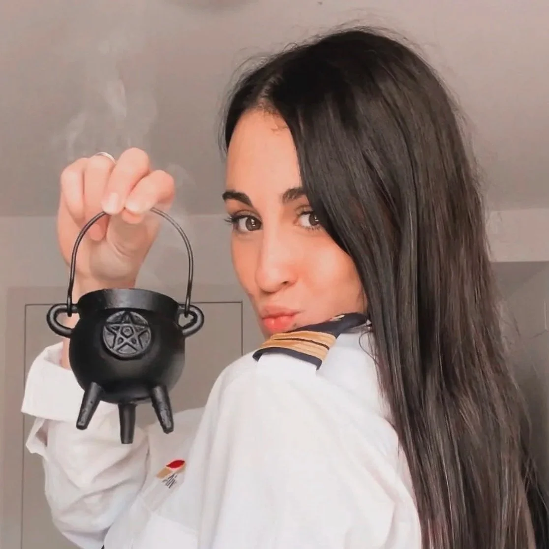 Woman with long dark hair holding a small black cauldron-shaped object emitting steam, looking at the camera.