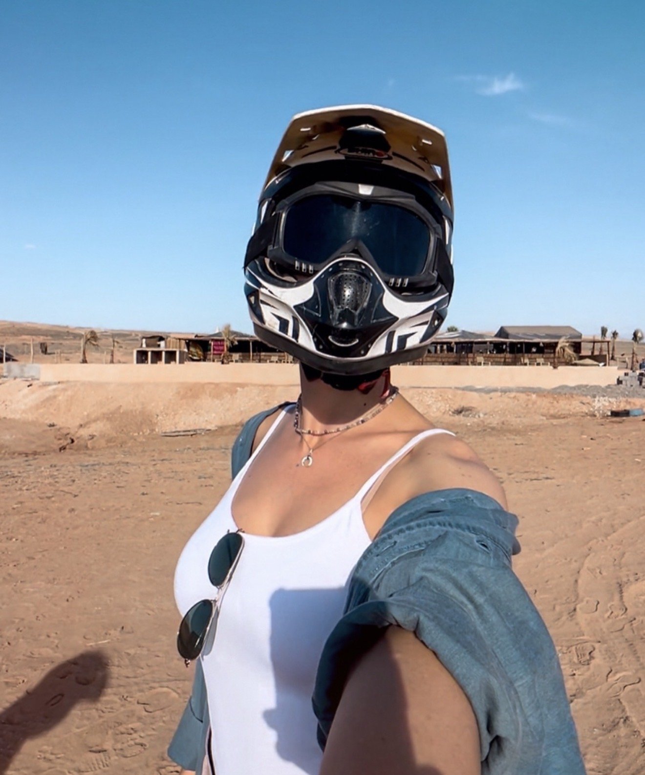 Person wearing a black and white helmet taking selfie in a desert with buildings and a clear blue sky in the background.