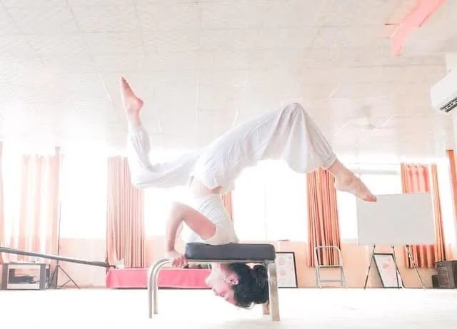 Person dressed in white performing a handstand on a padded obstacle in a sunlit room with pink curtains.