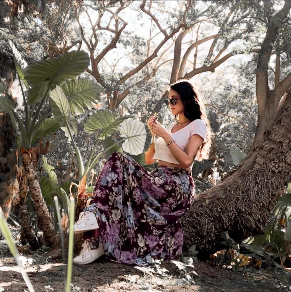 A woman with dark hair wearing sunglasses, a white crop top, and floral-patterned wide-leg pants sitting on a tree branch, holding a flower or plant, surrounded by large green leaves and trees.