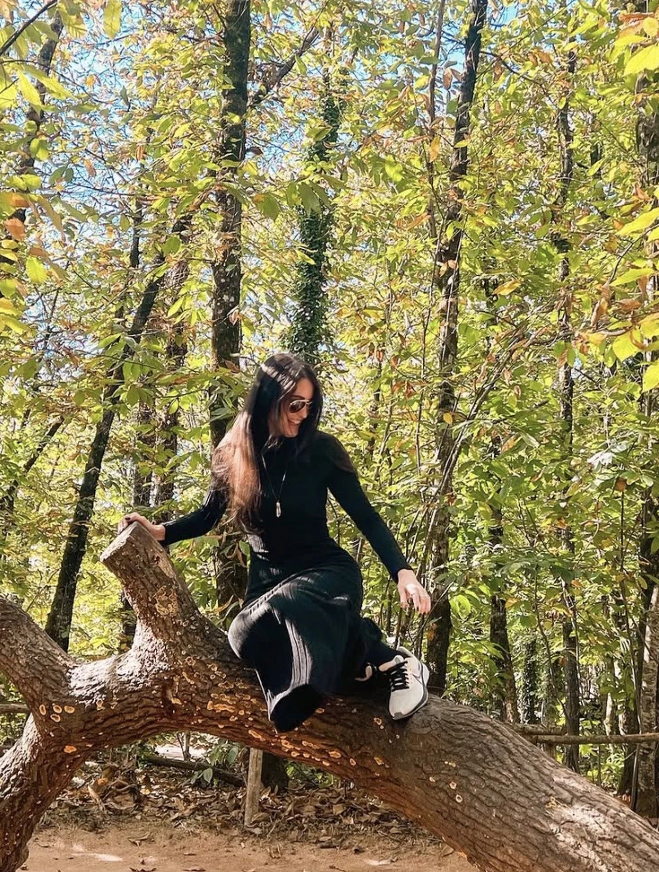 A young woman wearing sunglasses, black long sleeve shirt, black pants, and white sneakers sitting on a large tree branch in a leafy forest, smiling and looking down.