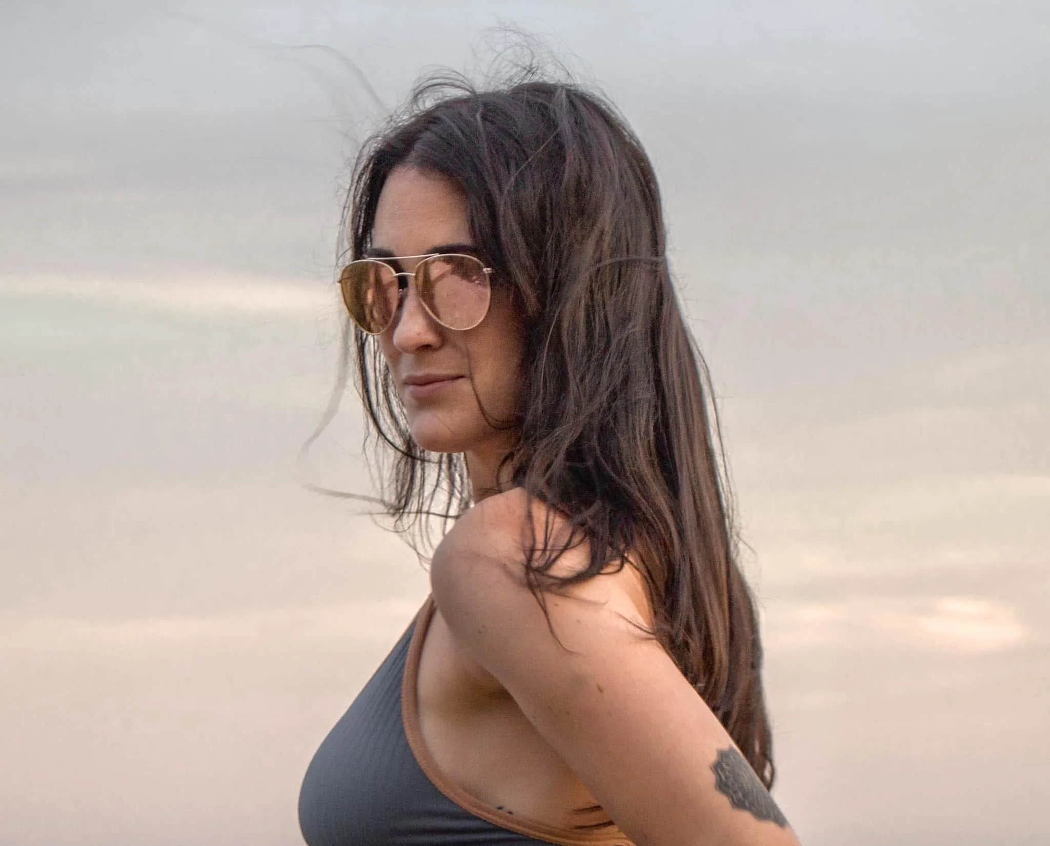 A woman with long dark hair wearing sunglasses and a black sports bra, standing outdoors facing sideways with her eyes closed, against a cloudy sky.