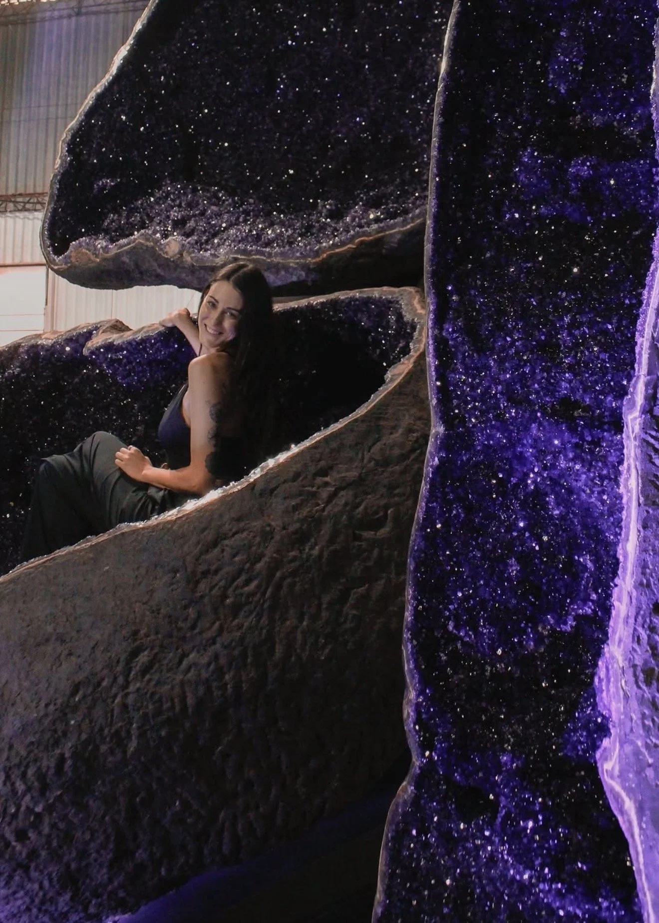 A woman sitting inside a large amethyst geode with purple crystalline interior, smiling at the camera.