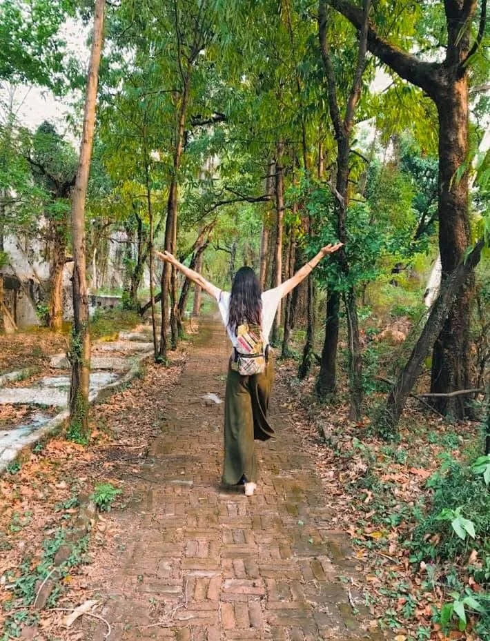 A woman walking on a brick path surrounded by green trees, with her arms raised in a joyful pose.