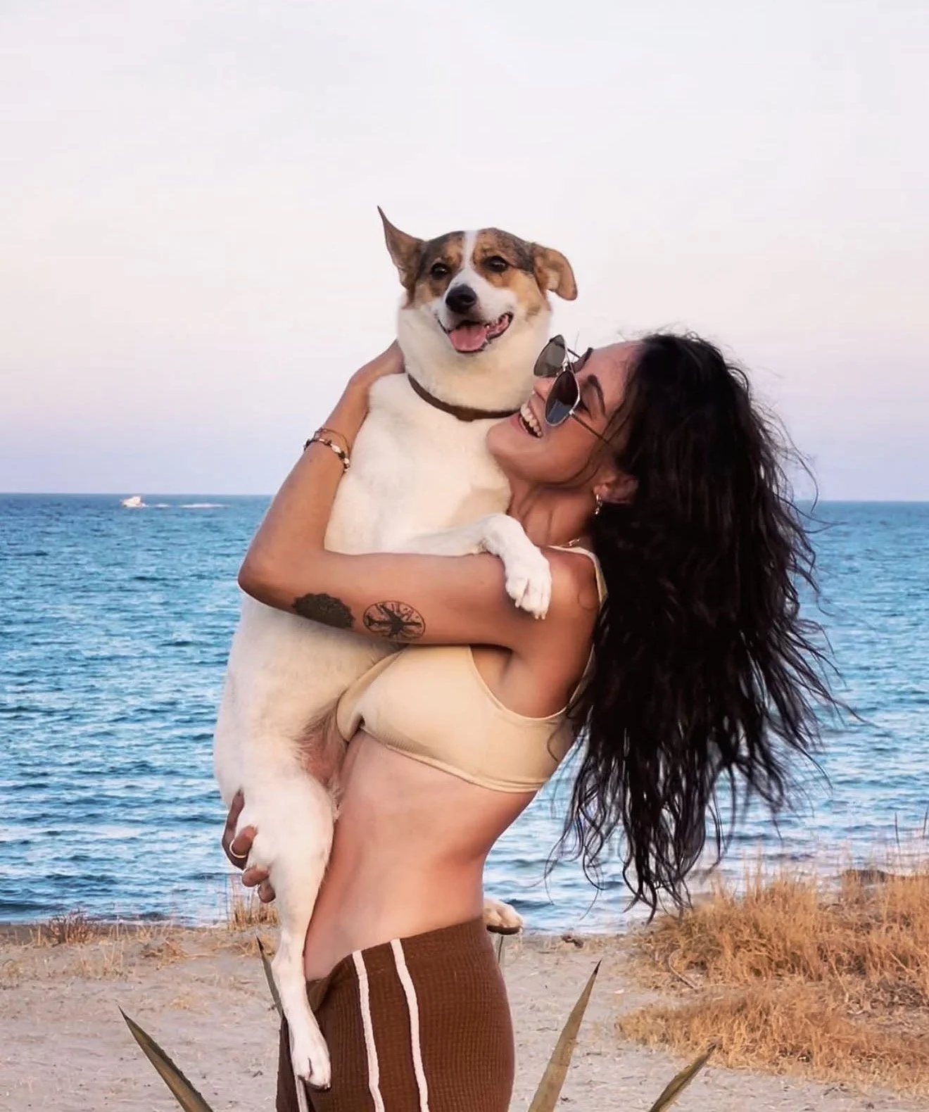 A woman with dark, curly hair, wearing sunglasses and a beige tank top, is smiling while holding a happy dog with a white and tan coat, against a backdrop of the ocean and a clear sky.