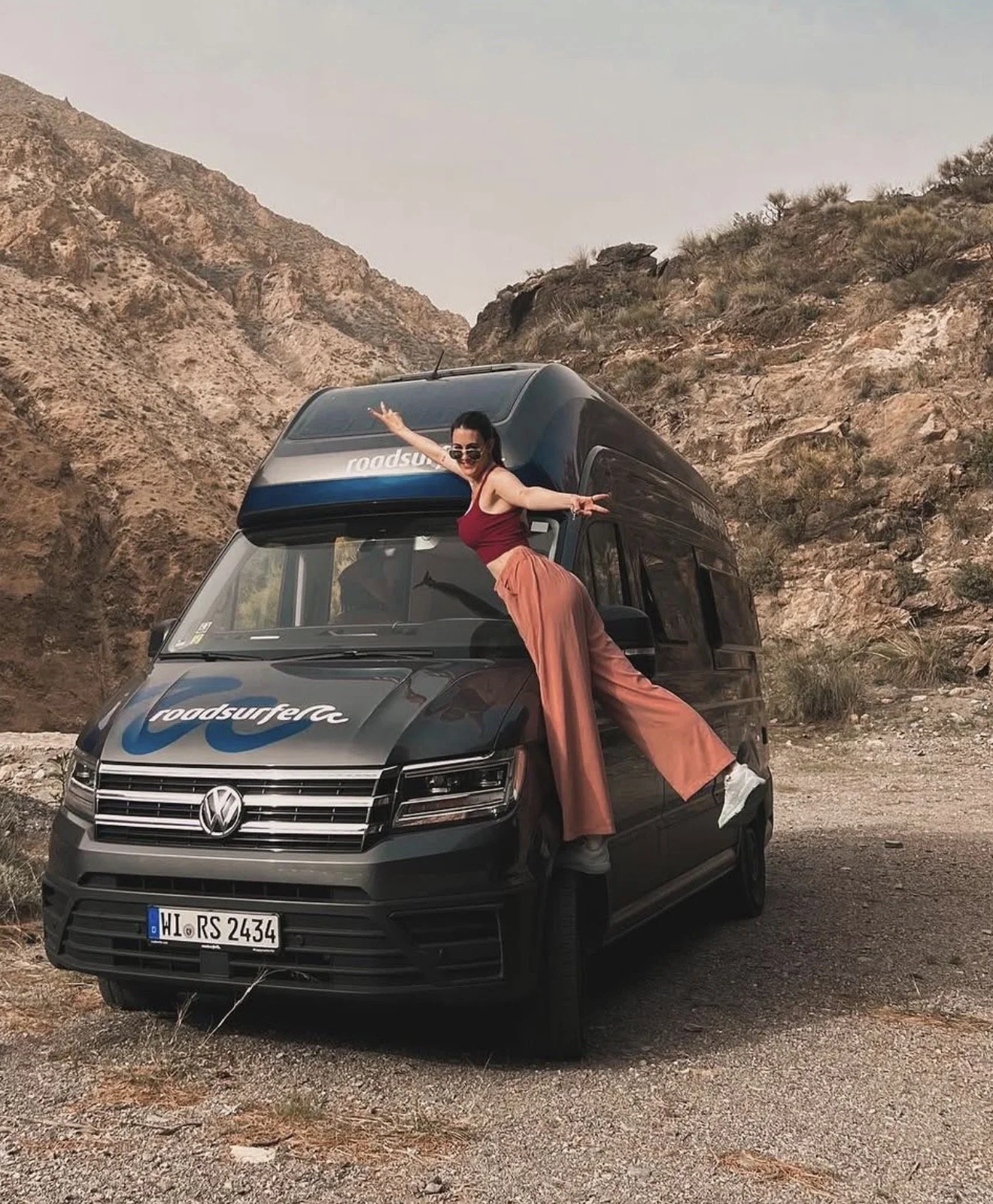 A woman with long dark hair, wearing sunglasses, a maroon tank top, and wide-legged orange pants, is standing on a black Volkswagen van with her arms outstretched. The van is parked on a gravel surface near rocky desert hills under an overcast sky, with the logo 'roadsurfer' on the front and a German license plate.