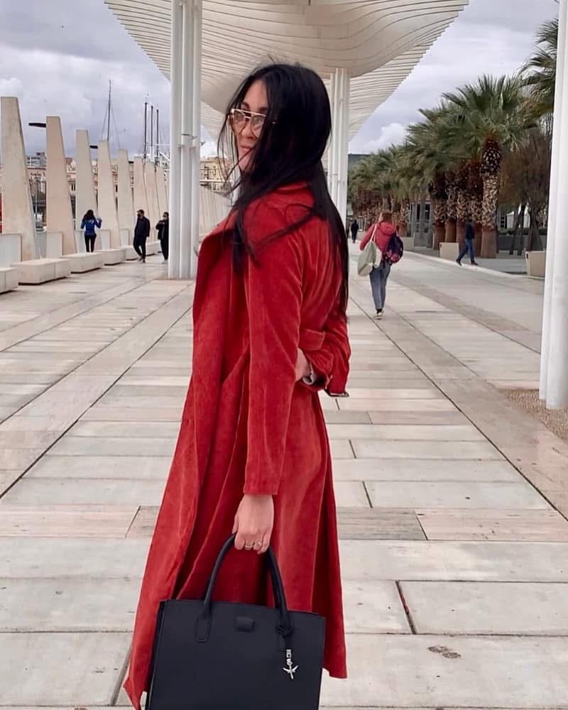 Woman in red coat with black handbag standing on a tiled promenade, with modern architecture and palm trees in the background.