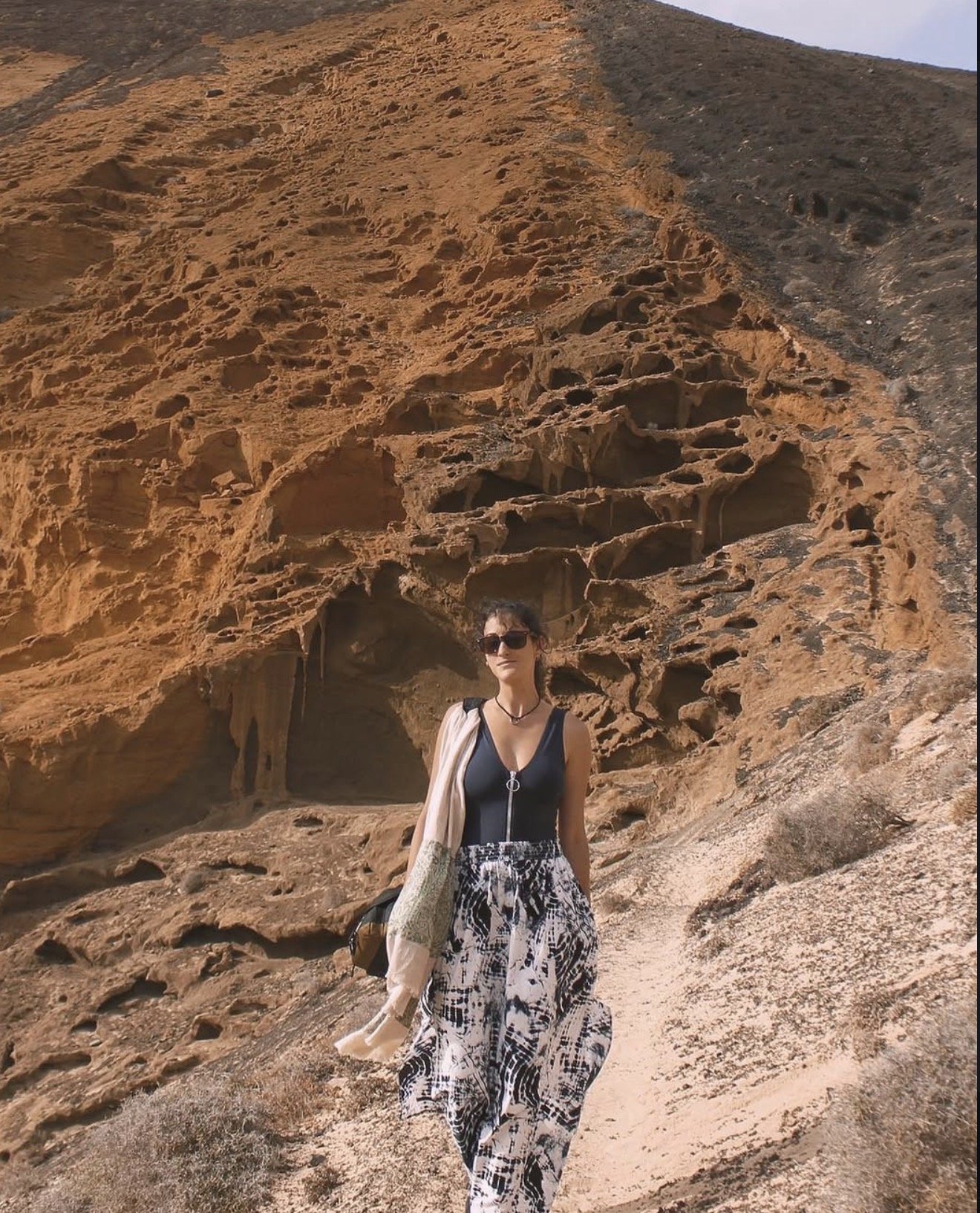 A woman wearing sunglasses, a black top, and patterned pants stands in front of a large, reddish rock formation with caves and irregular holes, under a blue sky.