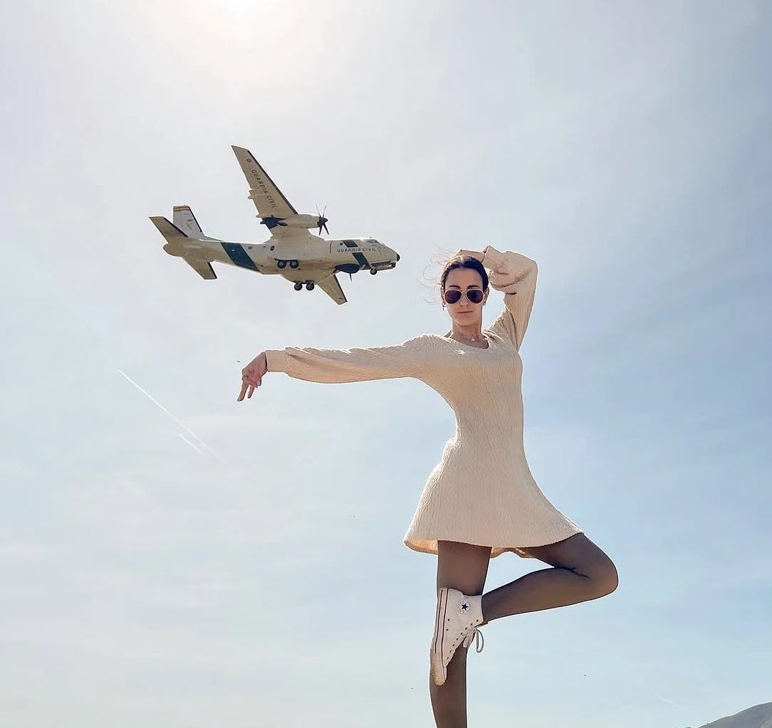 A woman wearing sunglasses and a beige dress posing outdoors against a cloudy sky with a toy airplane flying overhead.