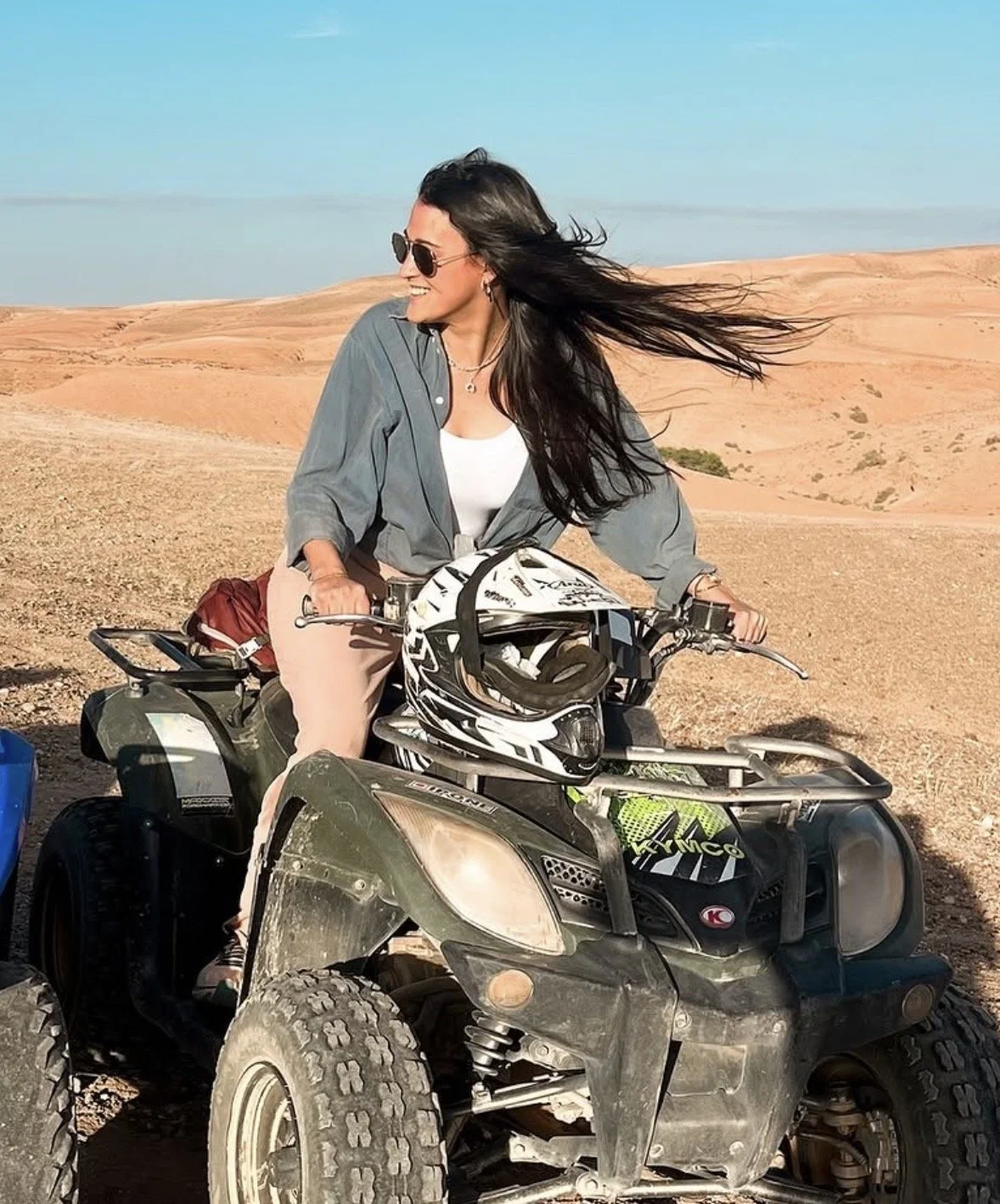 Woman with long dark hair wearing sunglasses and a gray shirt, riding a four-wheeler in a desert landscape with rolling sand dunes.