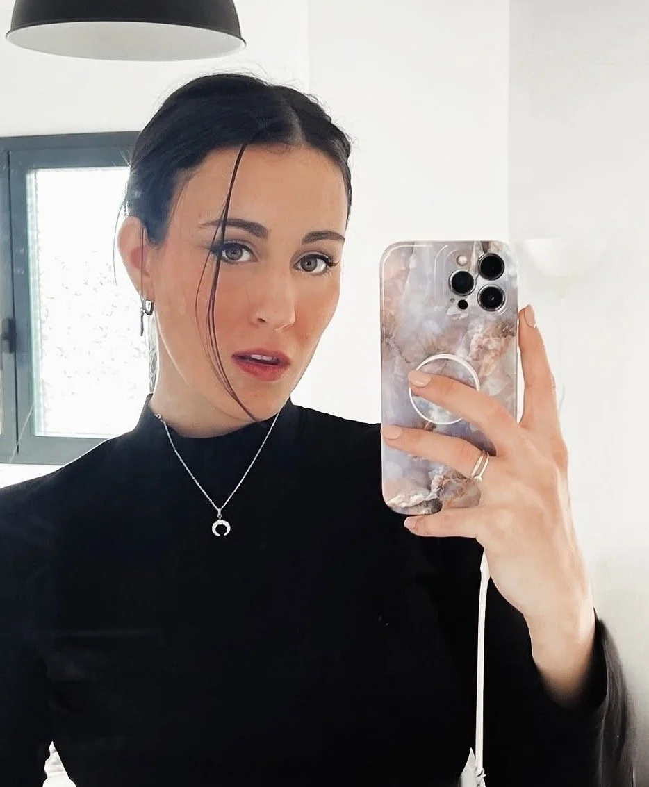 A woman taking a selfie with a smartphone mirror reflects her wearing a black top, silver jewelry, and has a hairstyle with a loose strand of hair. The background shows a window and a white wall.