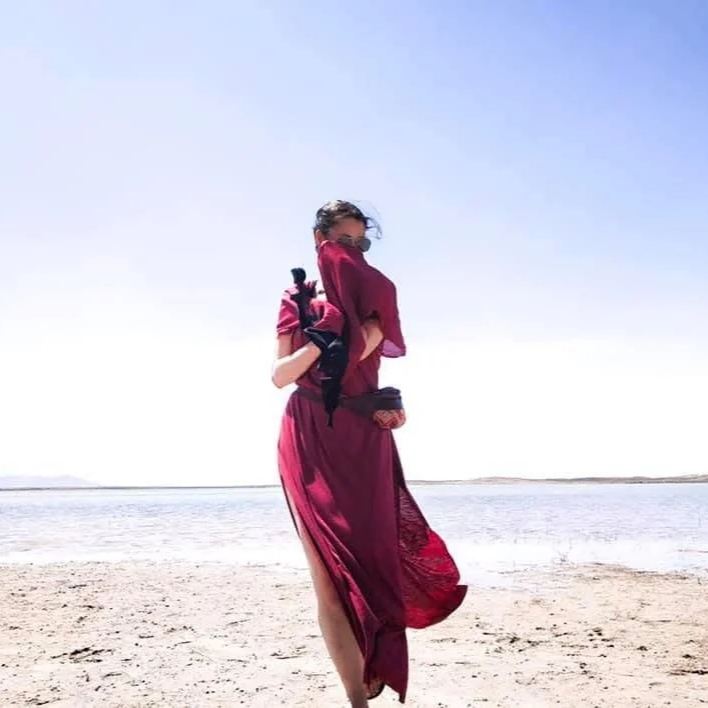 A woman wearing a burgundy dress and sunglasses stands on a beach, holding a black jacket over her shoulder with the ocean and a clear sky in the background.