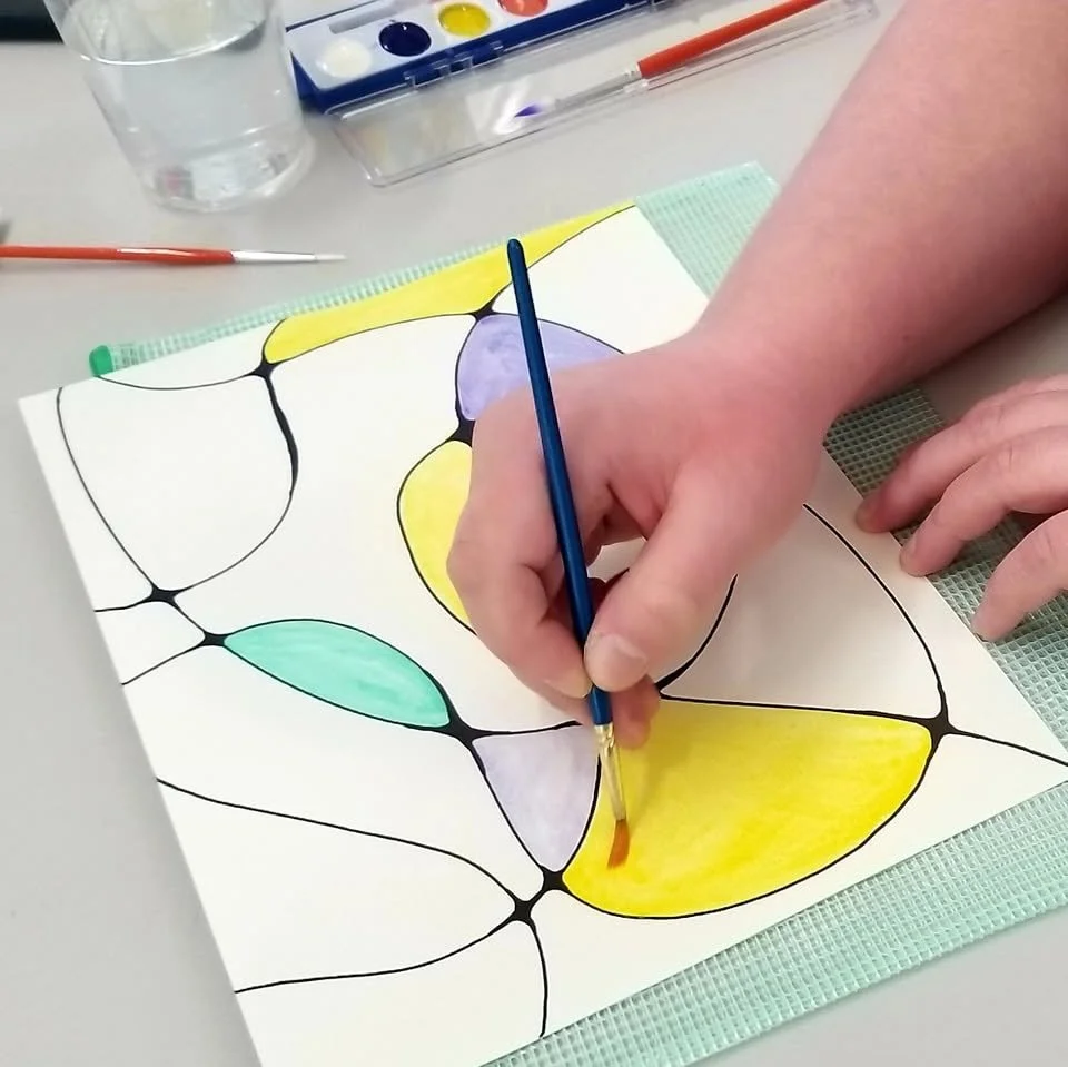 Person watercolor painting abstract design on paper, with various colored sections including yellow, purple, and teal, surrounded by black outlines.
