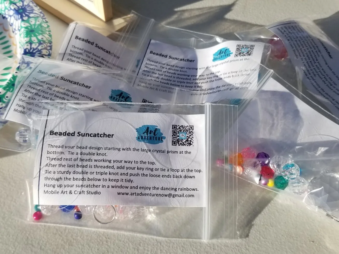 Clear plastic bags containing colorful beads with a printed instruction sheet for making beaded suncatchers, placed on a white surface.