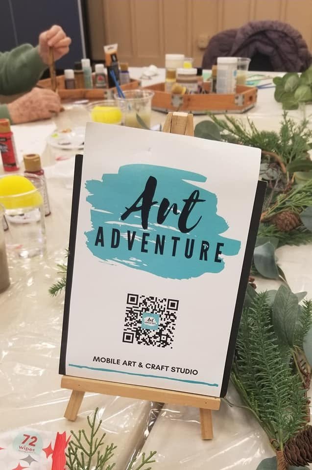 Sign advertising 'Art Adventure' mobile art and craft studio in the center of a craft table decorated with greenery, pinecones, and art supplies, with people working on art projects in the background.