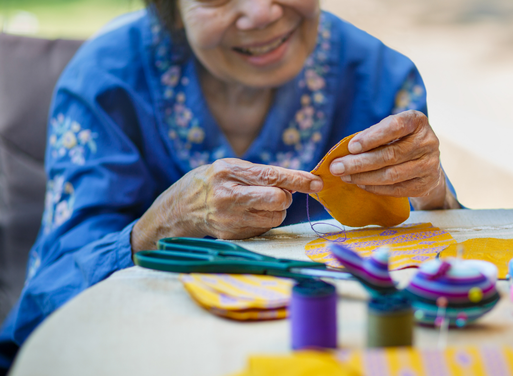 An elderly woman sewing yellow fabric with decorated embroidery, sitting at a table with sewing supplies.