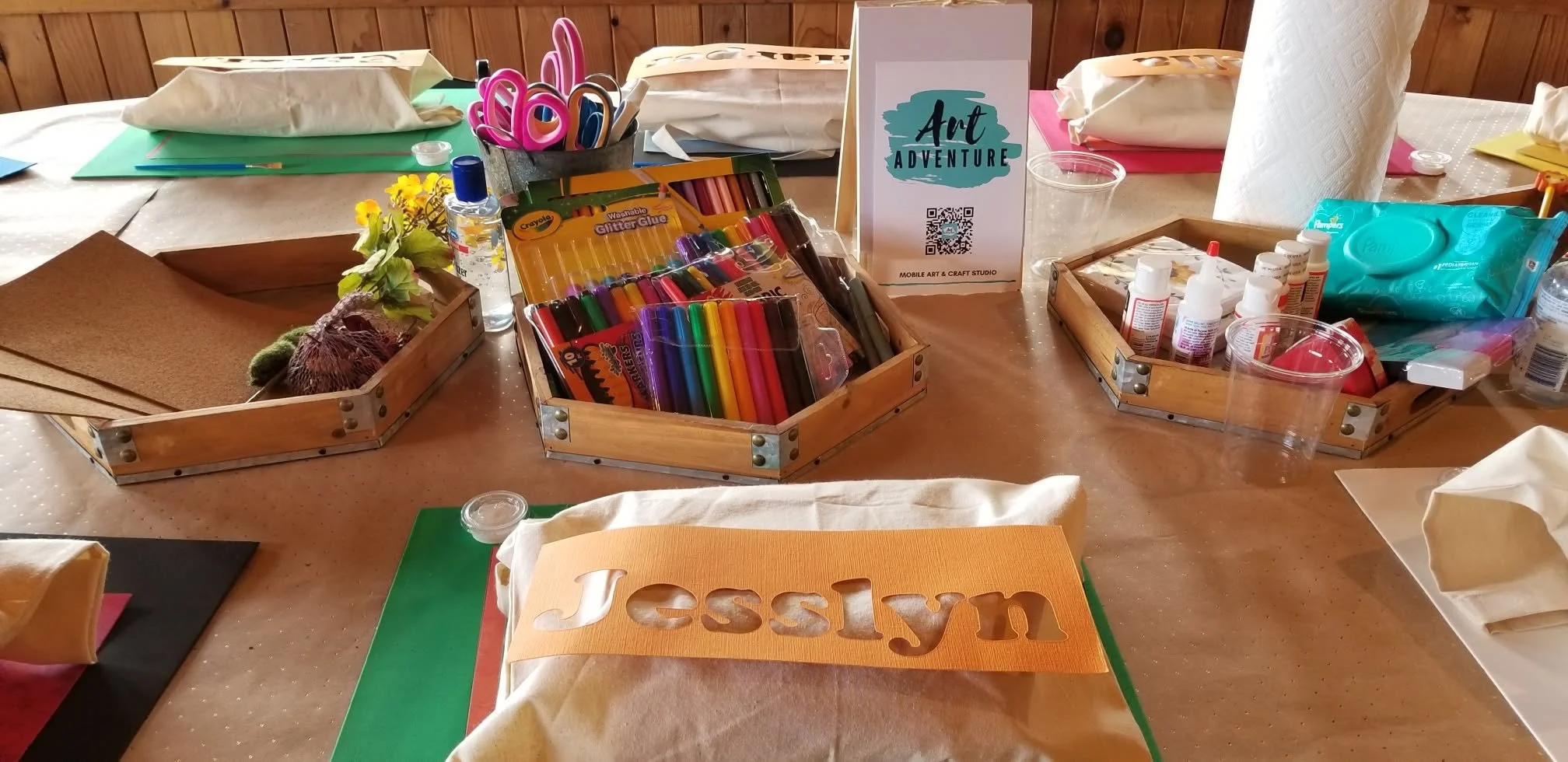 Table set up for arts and crafts with various supplies including markers, scissors, glue, tissues, and paper, with a sign that says 'Art Adventure' in the center.
