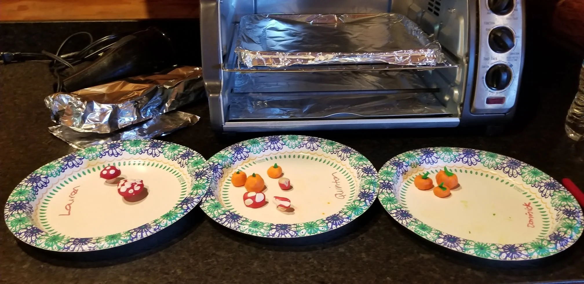 Three paper plates with colorful floral borders and names written on them, each with small pumpkin-shaped and mushroom-shaped decorations made of modeling clay. A mini oven and aluminum foil are visible in the background on a black countertop.