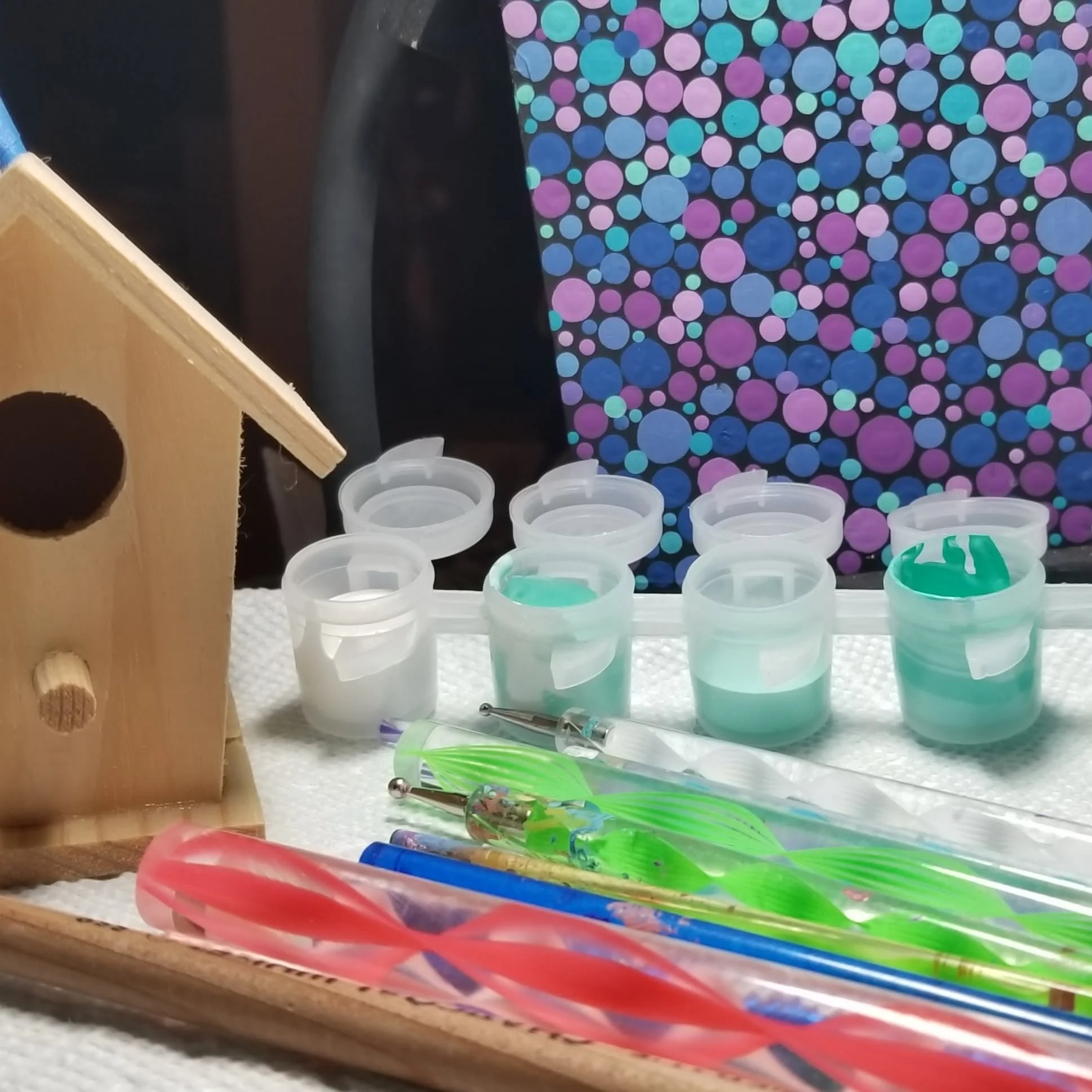 A collection of small plastic containers with teal-colored liquid, a wooden birdhouse with a large circular entrance, colorful swimbaits with feathered tails, and a green and purple fishing lure on a white textured surface.