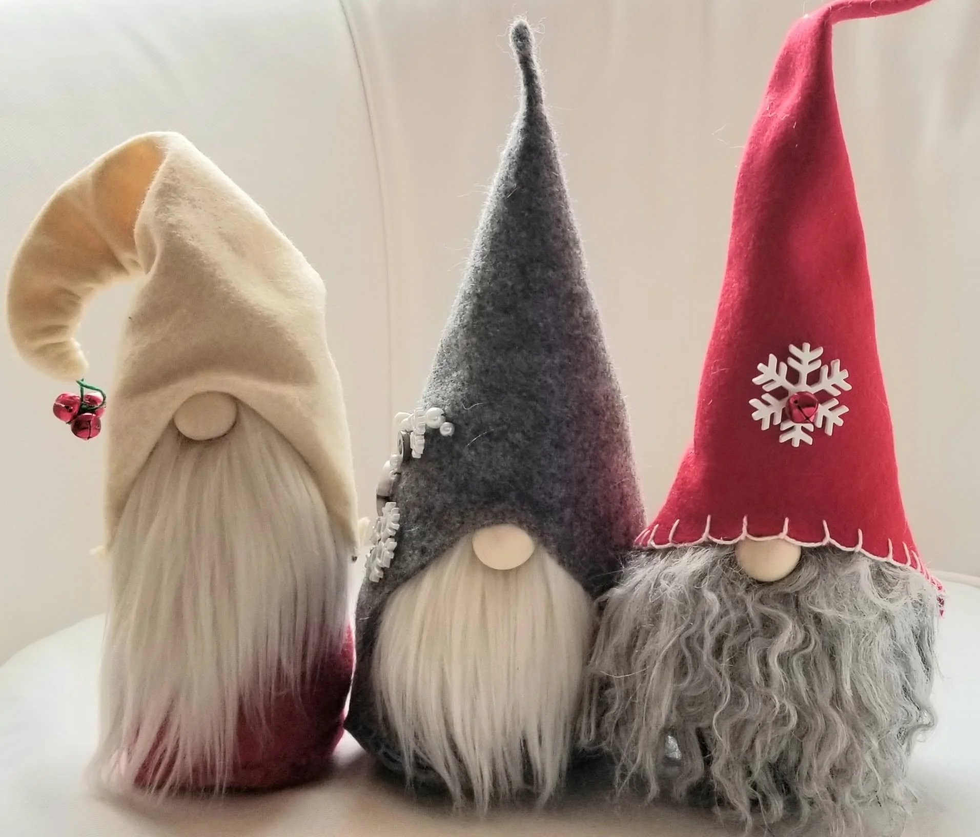 Three plush gnome decorations with long white beards, dressed in winter hats. The first gnome has a cream-colored hat with a bell and holly decoration. The second has a gray felt hat with white snowflake decorations. The third wears a red felt hat wi