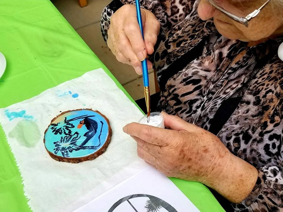 An elderly woman painting a snow leopard on a circular wooden slice surrounded by a green tablecloth.
