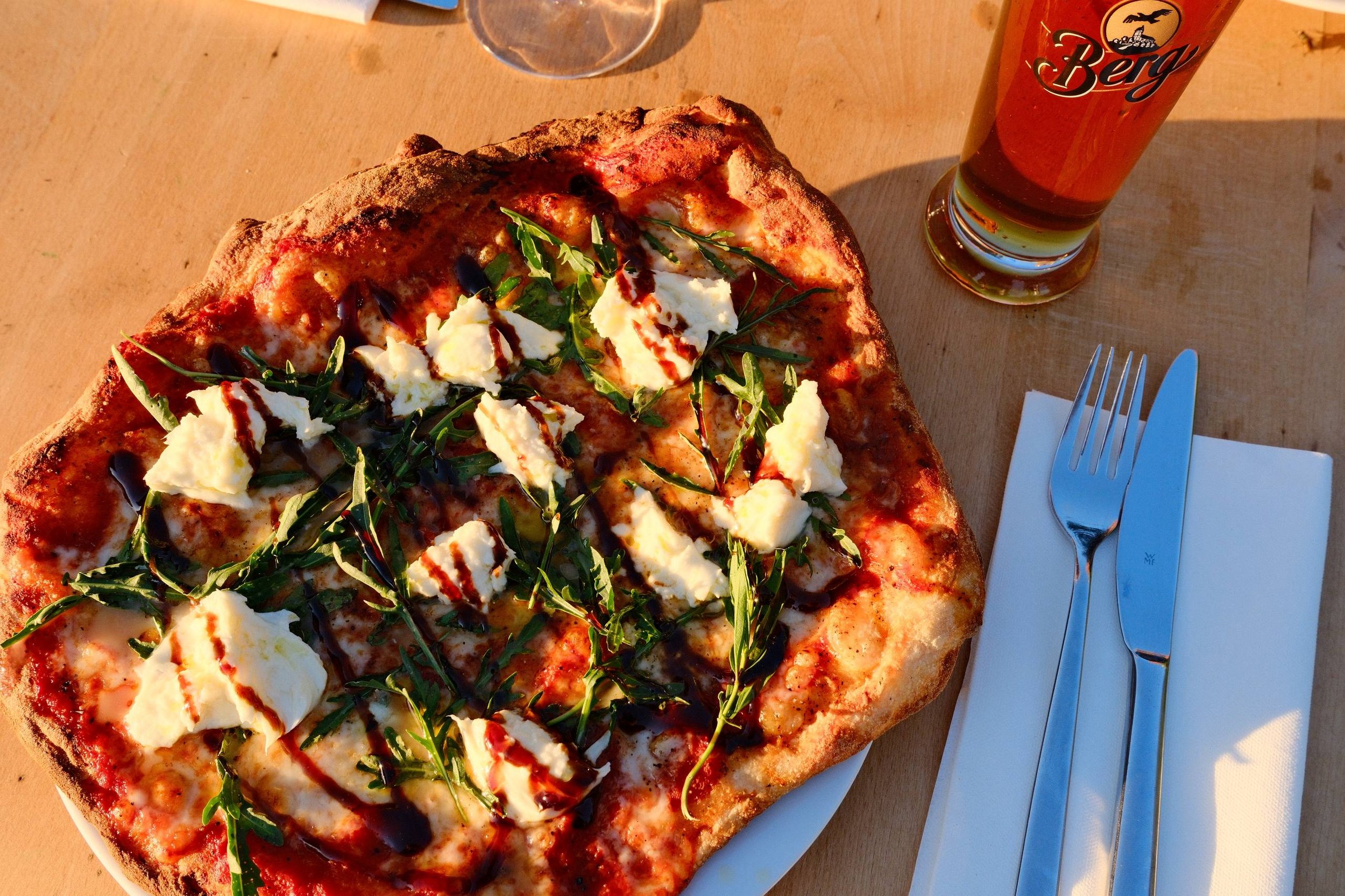 Pizza mit Tomatensauce, Mozzarella, Rucola und Balsamico auf einem weißen Teller, daneben ein Glas Wasser, auf einem Holztisch.