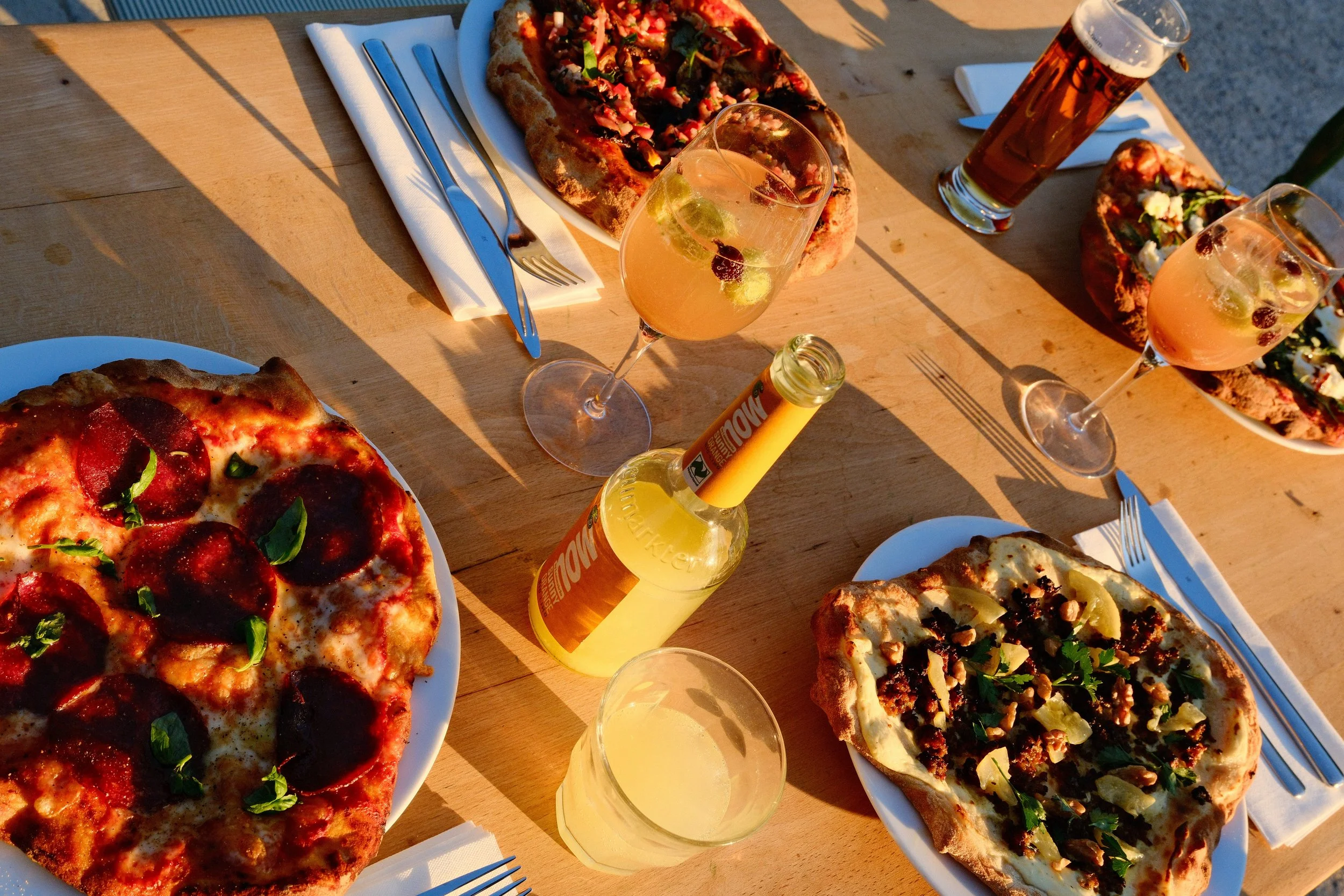 Vier Pizzen auf einem Holztisch, je eine mit verschiedenen Belägen wie Salami, Gemüse und Käse. Zwei Gläser mit Bier, eines mit Limonade und Zitronenscheiben. Besteck und Papiertücher auf dem Tisch.