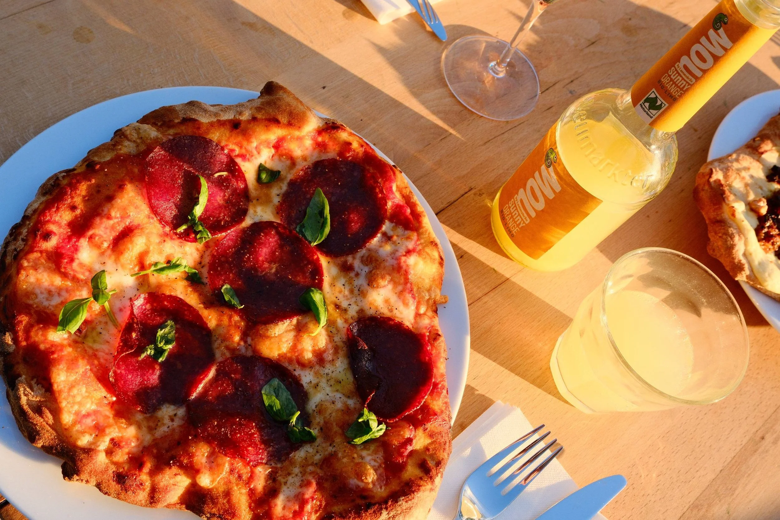 Eine Portion Pizza mit Salami und Basilikum auf einem weißen Teller, daneben ein Glas mit gelber Getränk und eine Flasche mit einem ähnlichen Getränk. Im Hintergrund befinden sich Gabel, Messer und eine leere Tasse auf einem Holztisch.