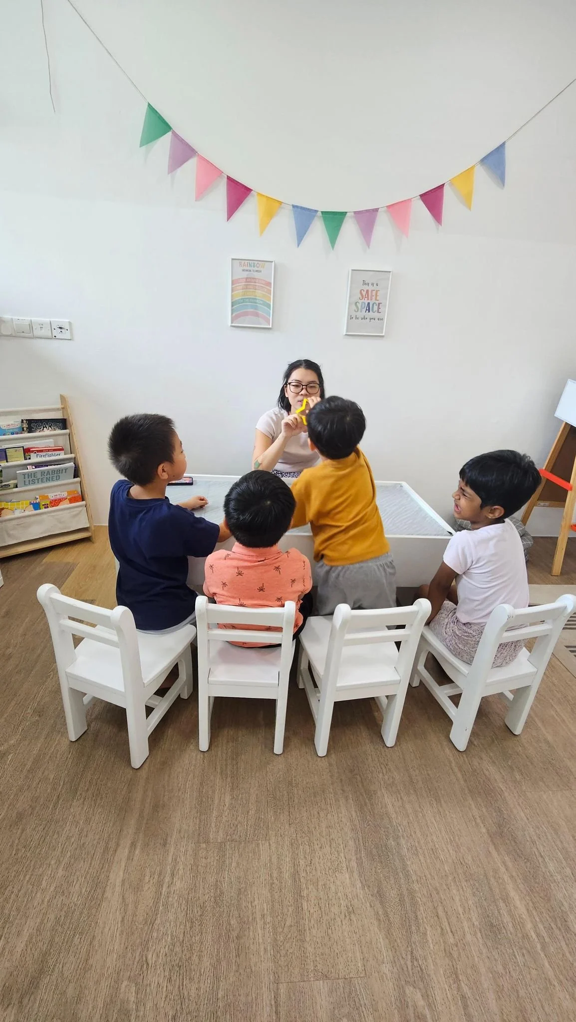 A woman conducts a group activity with five children in a room decorated with colorful bunting and framed posters, sitting around a white table.