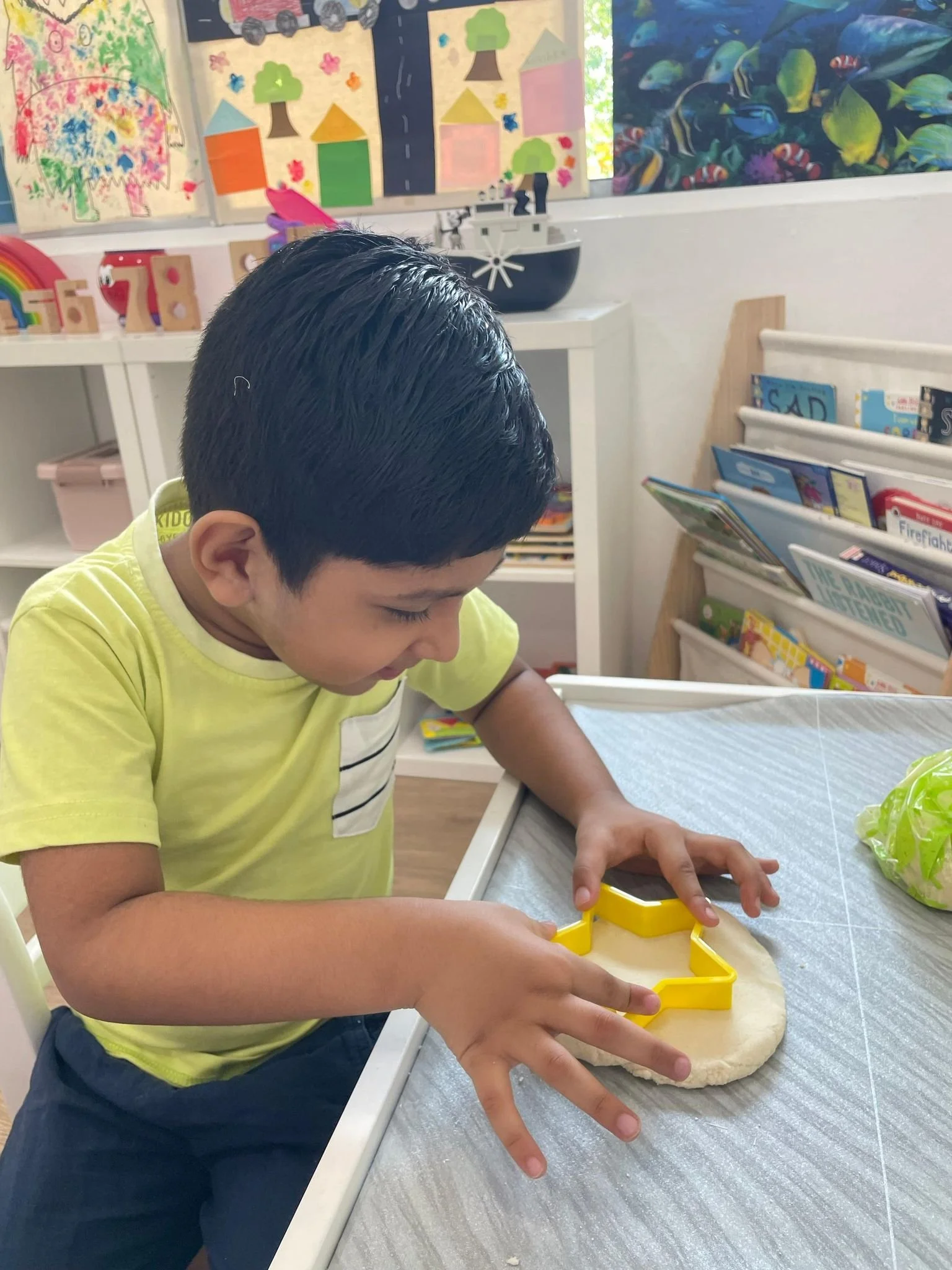 A young boy with short dark hair is using a yellow cookie cutter to cut dough on a table in a classroom decorated with colorful children's artwork on the walls and shelves filled with books and toys.