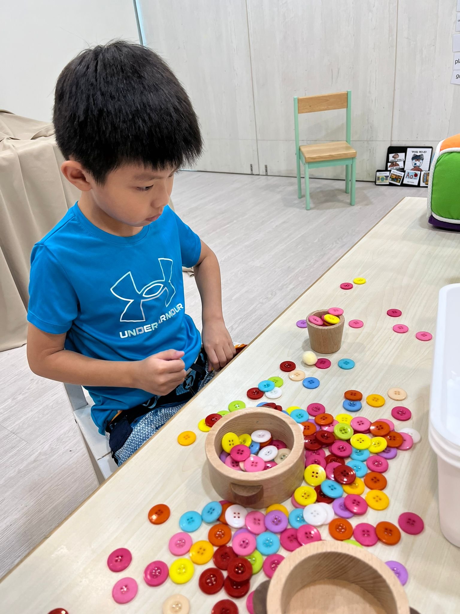 A young boy in a blue Under Armour shirt is sitting at a table with colorful buttons scattered across the surface. There are two small wooden bowls filled with buttons, and some buttons are spilled onto the table. In the background, there is a green and wooden chair, some artwork on the wall, and a colorful storage cube.
