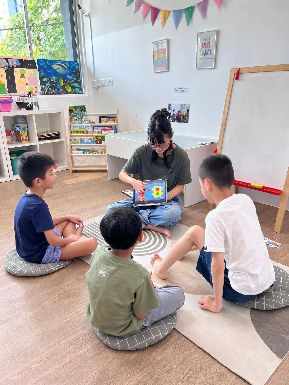 A woman sitting on the floor in a classroom with four children, engaging in a learning activity with a tablet device, while the children sit on cushions on a patterned rug. The classroom has colorful decorations, books, and artwork on the walls.