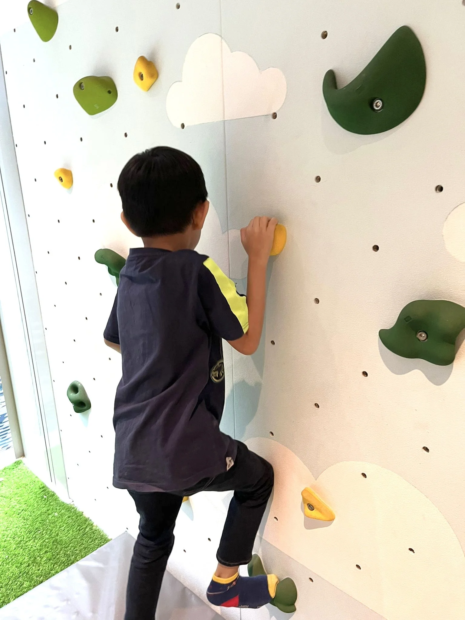 A boy climbing an indoor rock climbing wall with colorful holds and cloud-shaped decorations.