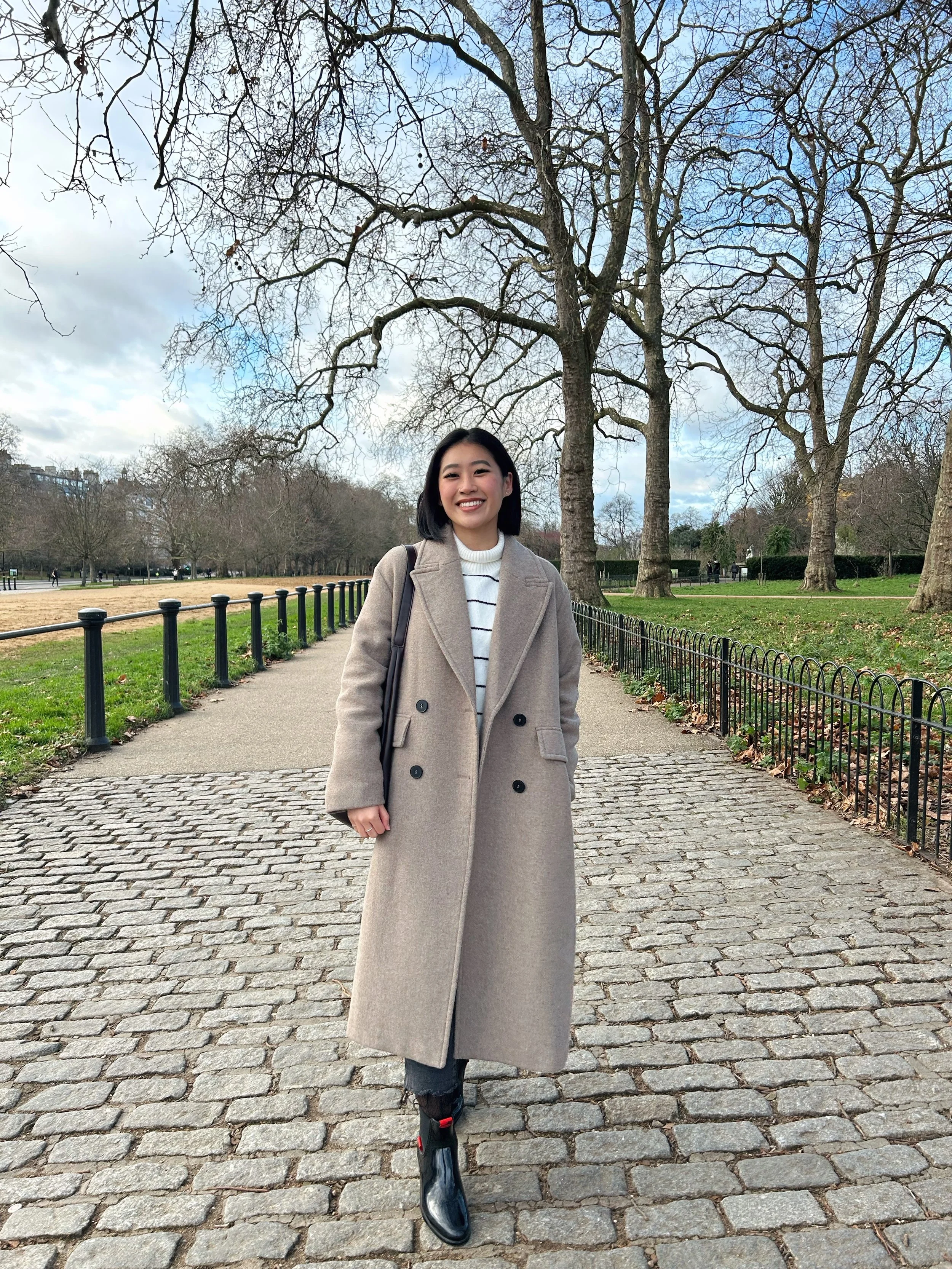 A woman with black hair smiling, wearing a beige coat and striped sweater, standing on a cobblestone path in a park with leafless trees and a fence in the background.