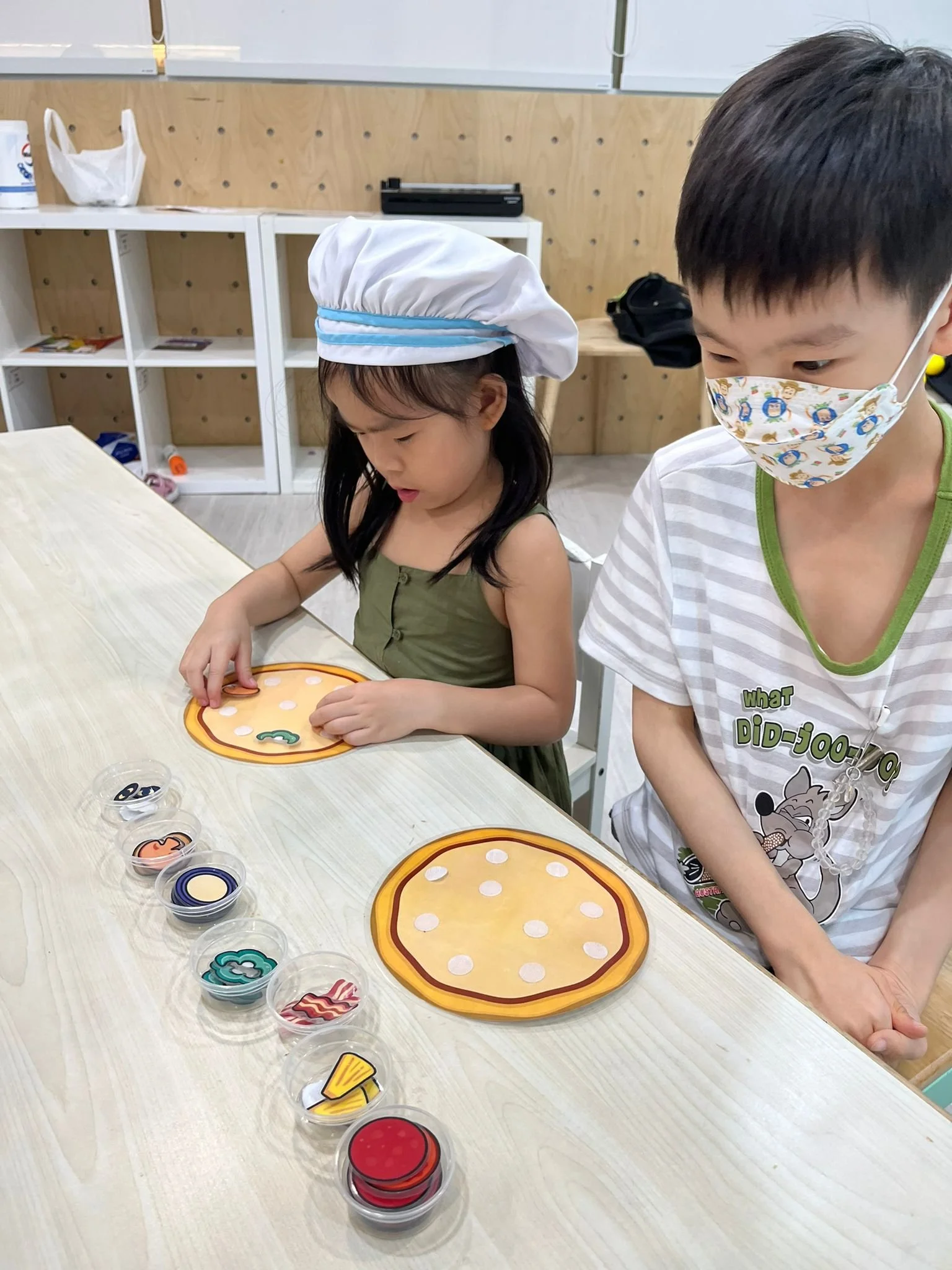 Children playing a tabletop game with small, colored obstacles and a circular game board, with containers of game pieces on a light-colored table in a classroom or activity room.