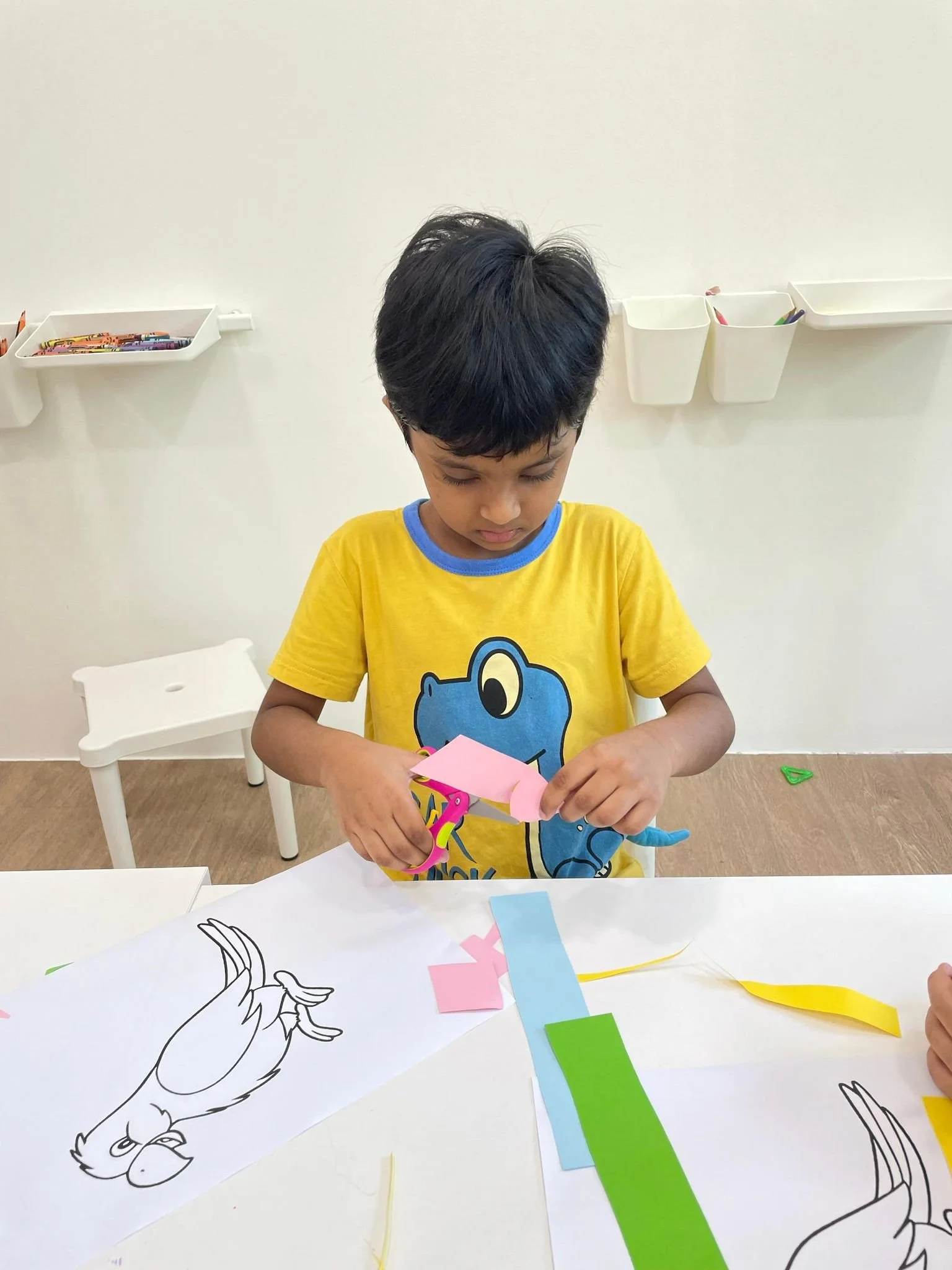 A young boy with dark hair wearing a yellow T-shirt with a cartoon character is cutting colored paper with scissors at a table with arts and crafts supplies, including a coloring sheet of an illustrated rooster.