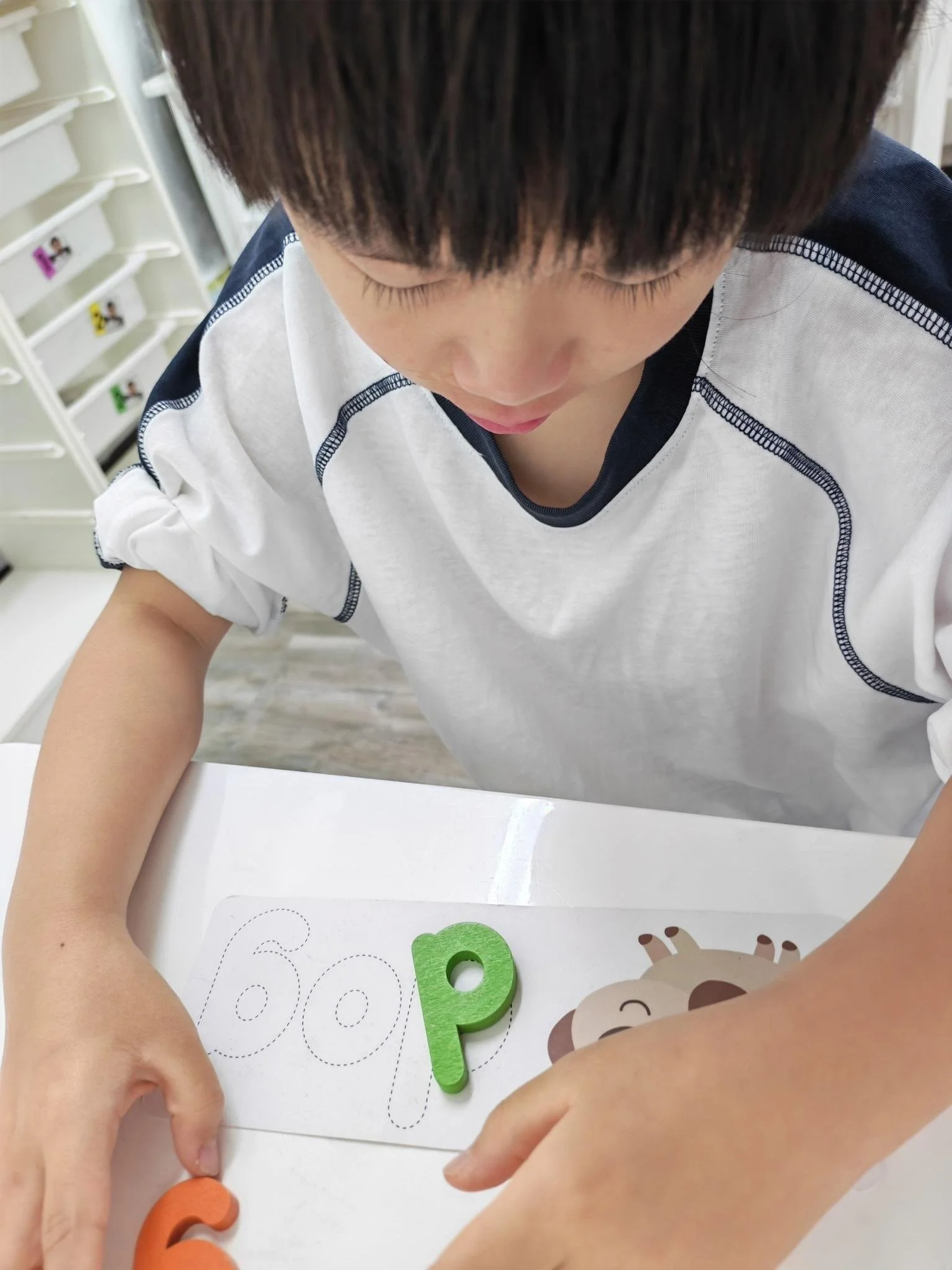 A child with short dark hair playing with foam letter cutouts and a workbook with dotted lines on a white table. An adult's arm is visible guiding the child's hand.