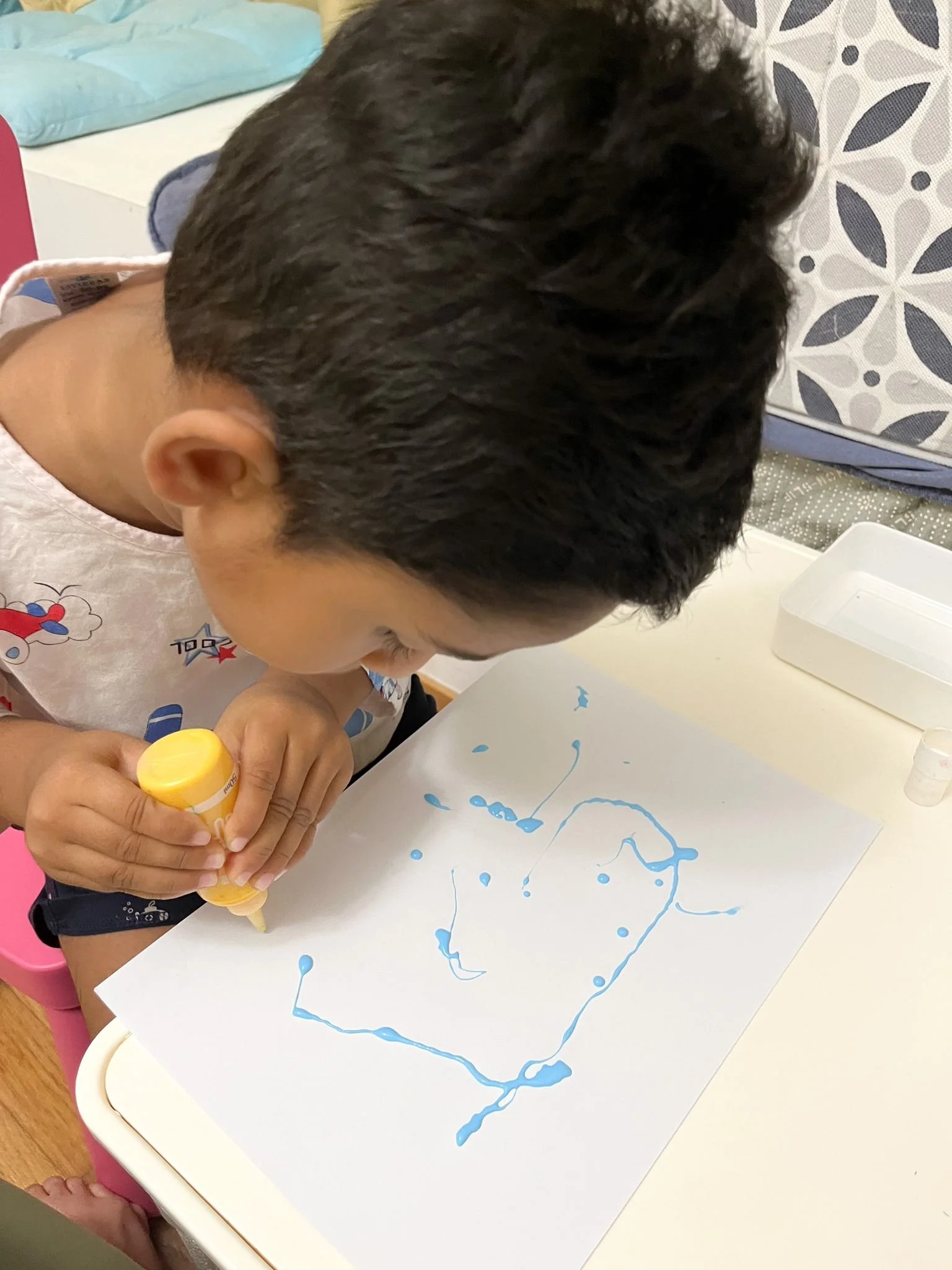 A young boy with black hair is drawing on a white sheet of paper with blue paint or ink, creating a line drawing or abstract design. The boy is wearing a sleeveless shirt and is focused on his artwork.