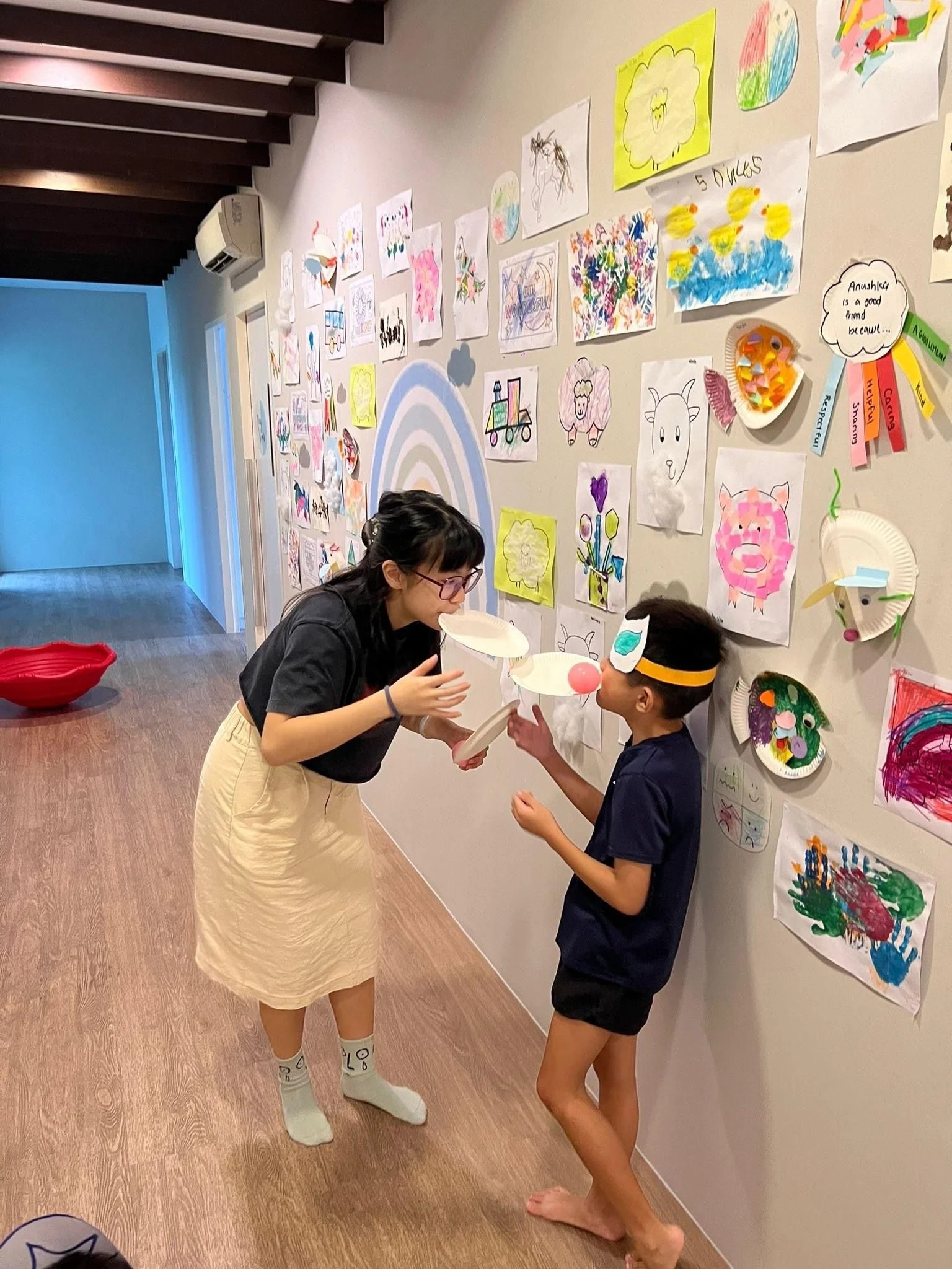 A woman and a young boy are engaging in a playful activity in a colorful art-filled room. The woman is wearing a yellow skirt and white socks, and the boy is wearing dark shorts and a blindfold while holding a plate. The wall behind them is decorated with numerous children's drawings and crafts, including animals and rainbows.