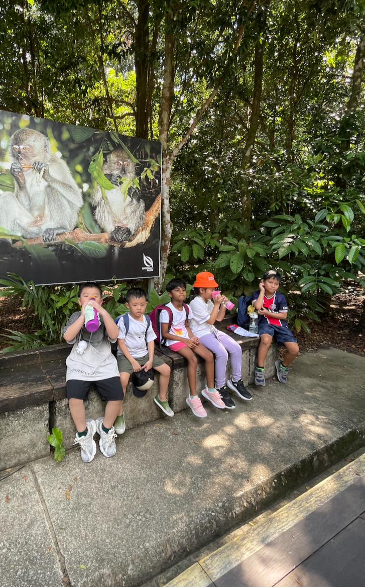 Five children sitting on a bench in a lush, green outdoor setting with a large sign featuring images of monkeys.